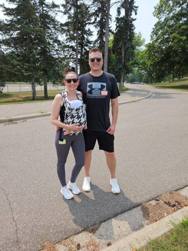 Couple walking with a baby carrier on a paved path, trees in the background on a sunny day.