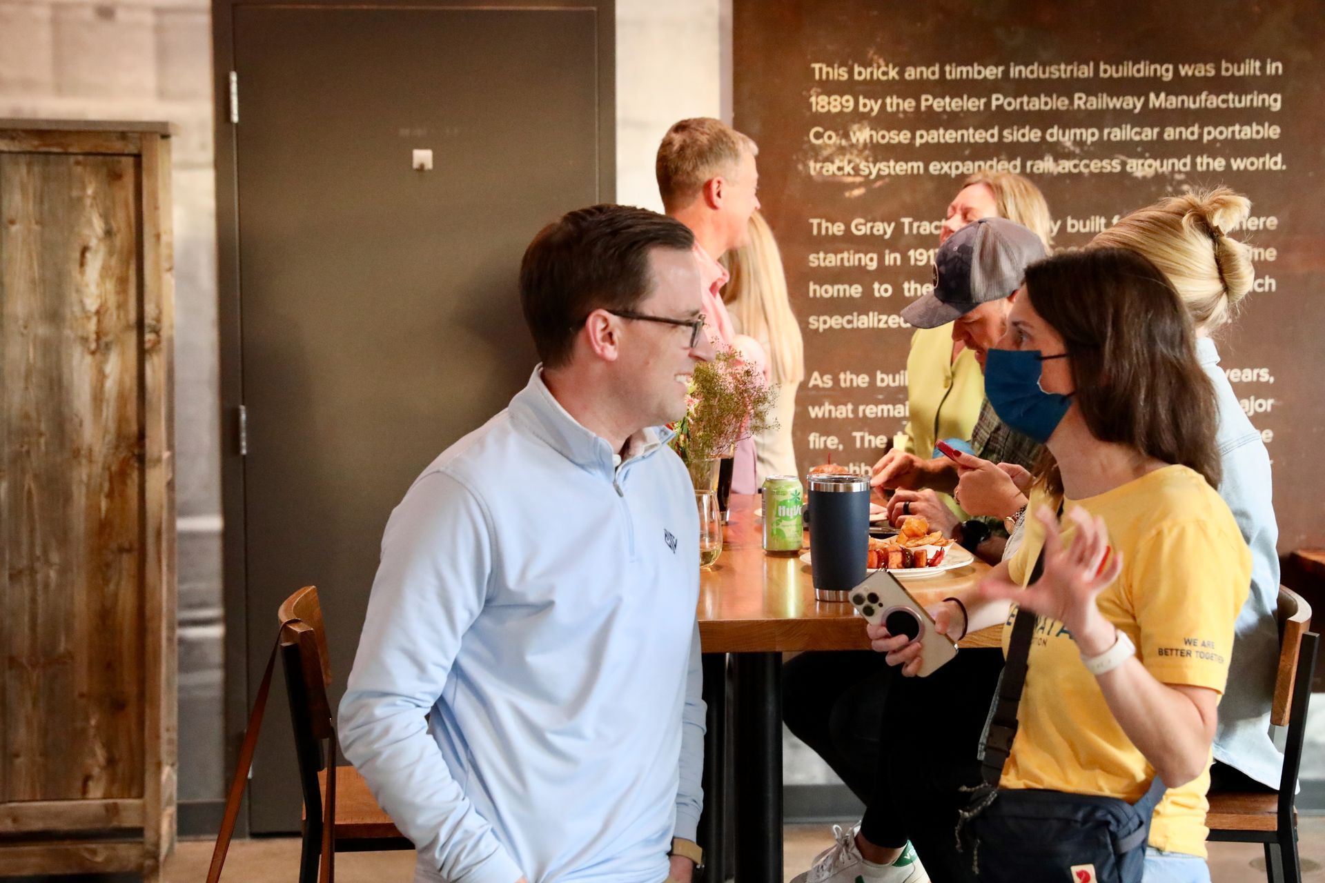 People in an office setting. A man in a light blue shirt talks with a woman in yellow. Others chat around a table.