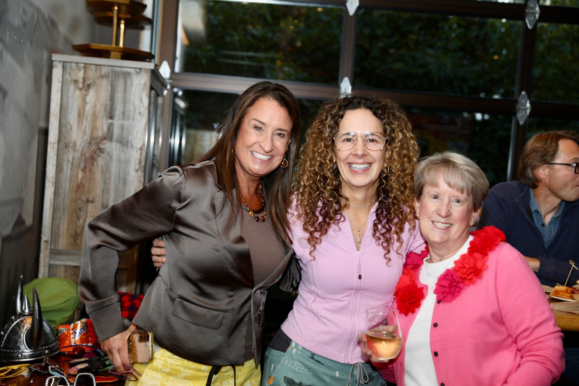 Three women smiling, embracing. One in brown, one in pink, and one with curly hair. They are indoors.