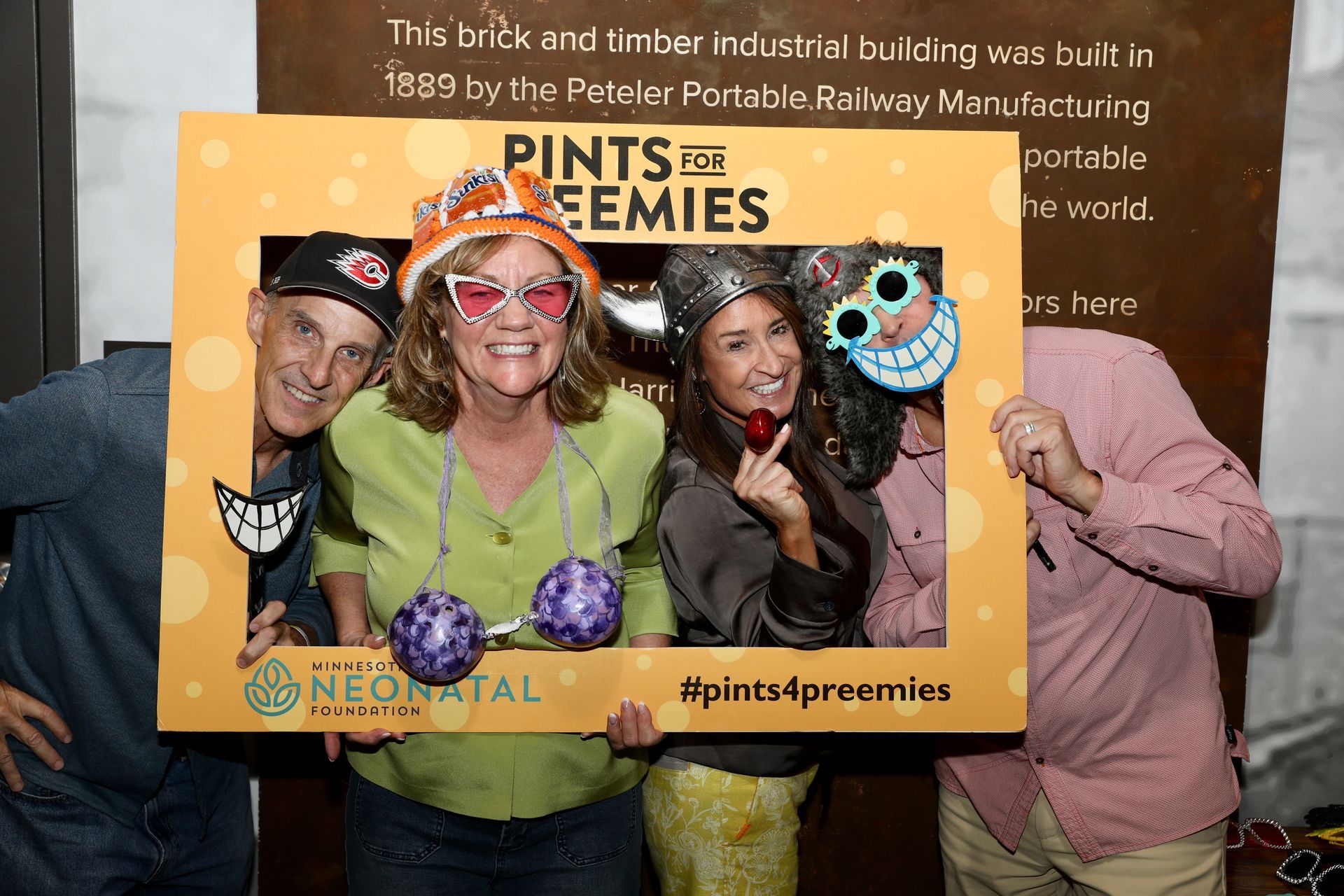 Four people posing behind a photo booth frame, decorated with props, at a Neonatal event.