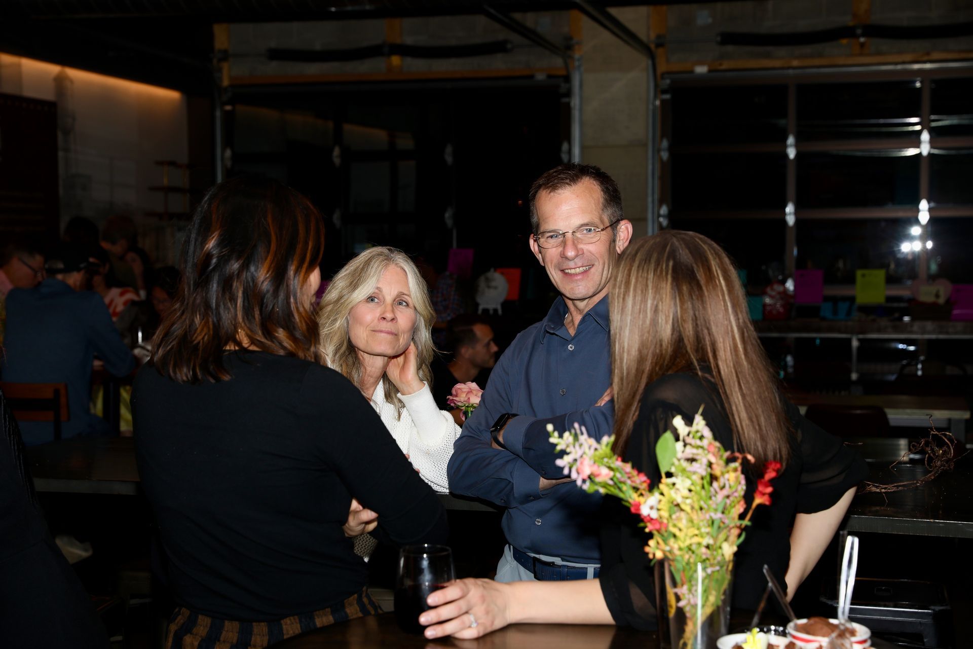 People in a dimly lit bar, conversing near a counter with a flower arrangement.