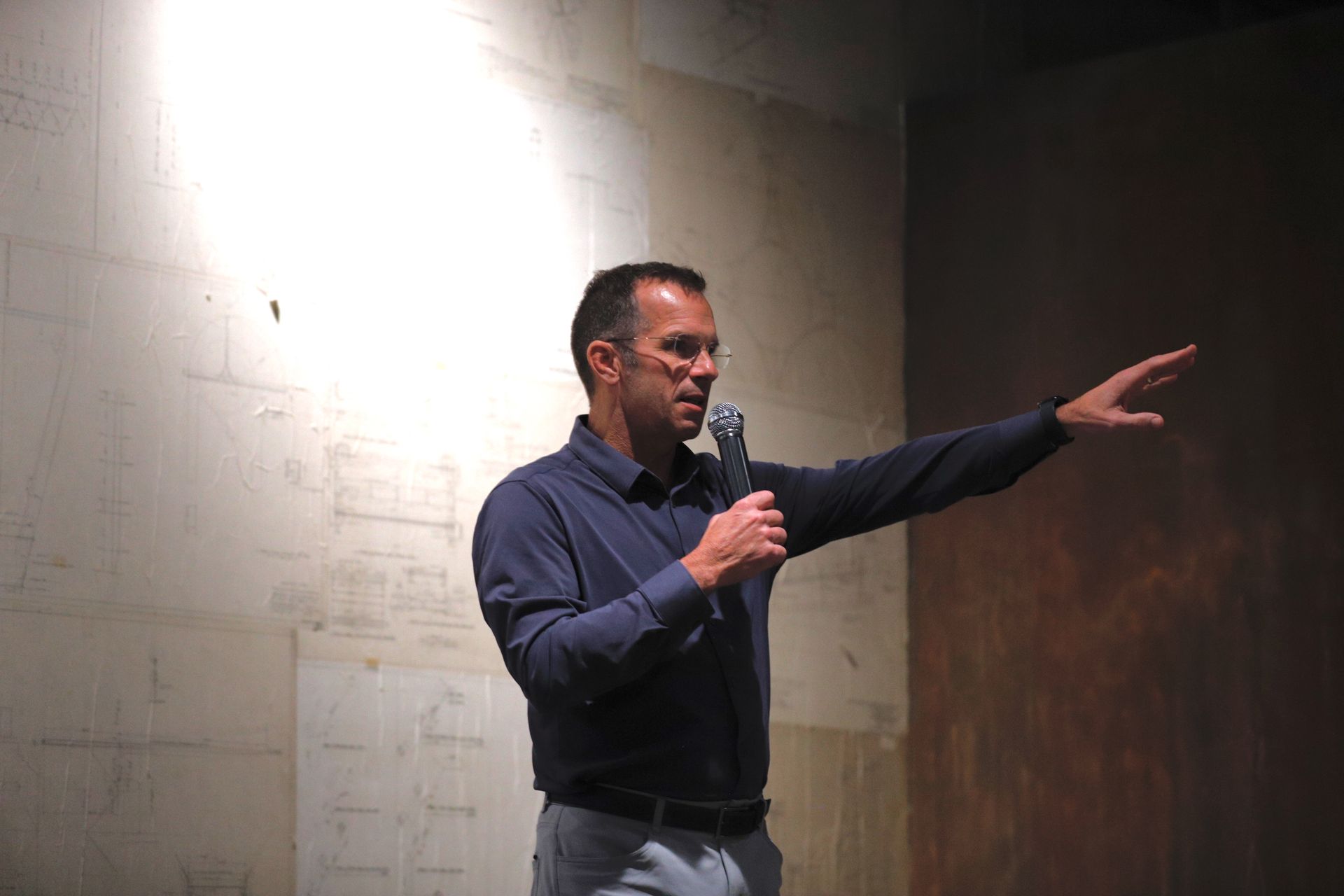 Man in blue shirt gestures right while holding a microphone, speaking. Stands near a wall in a dimly lit setting.