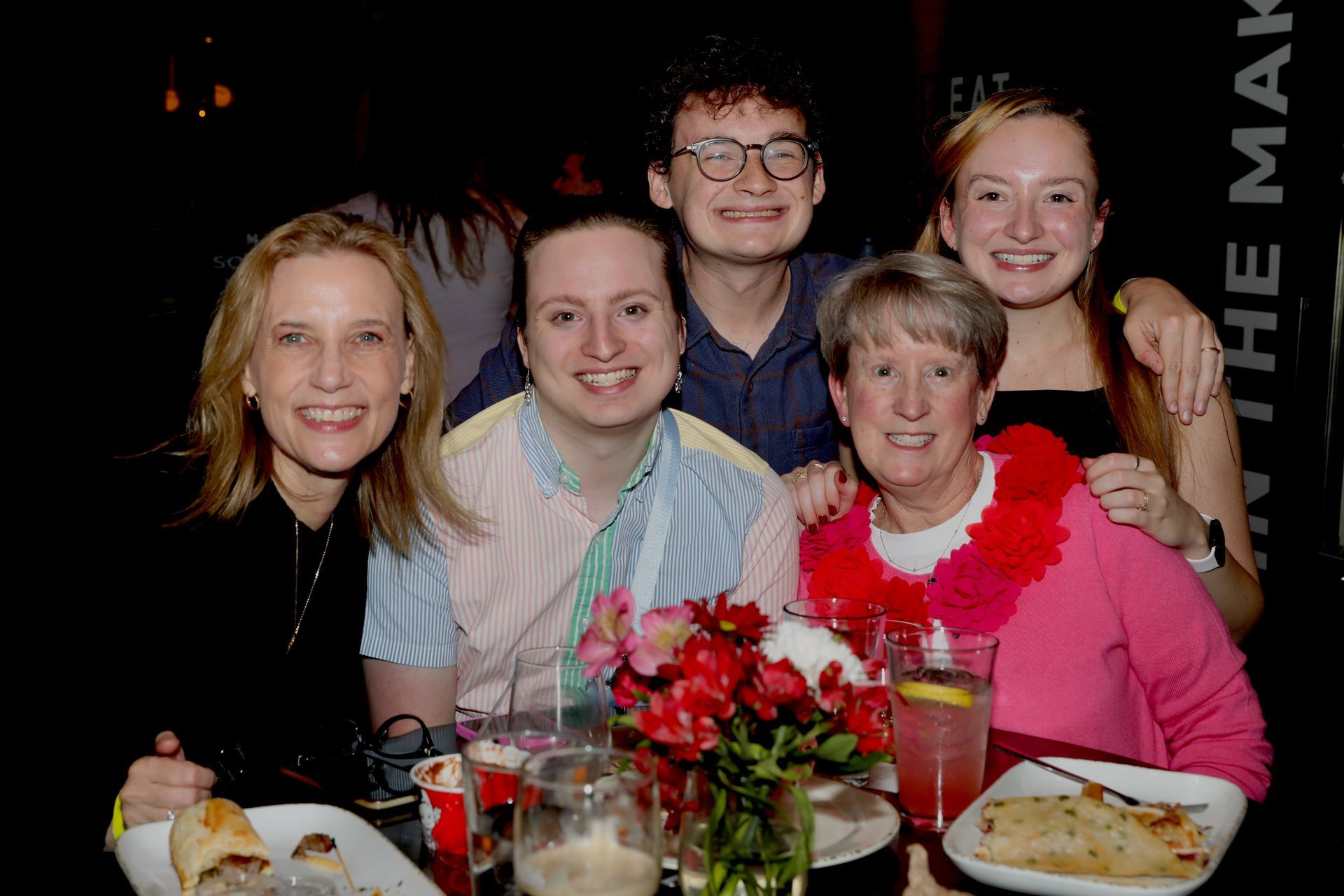 Five people smiling at a table with food and drinks; one wears a pink lei. Dimly lit setting.