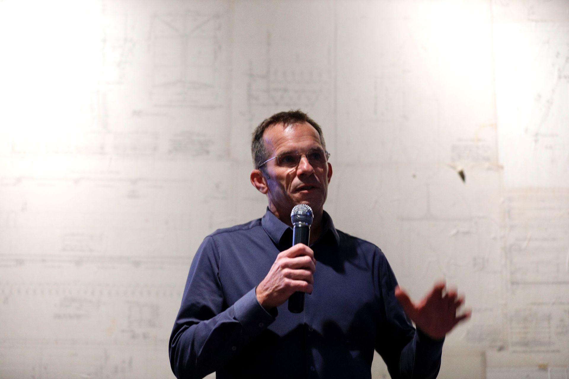 Man speaking into a microphone, gesturing with his hands. Against a white background with faint lines.