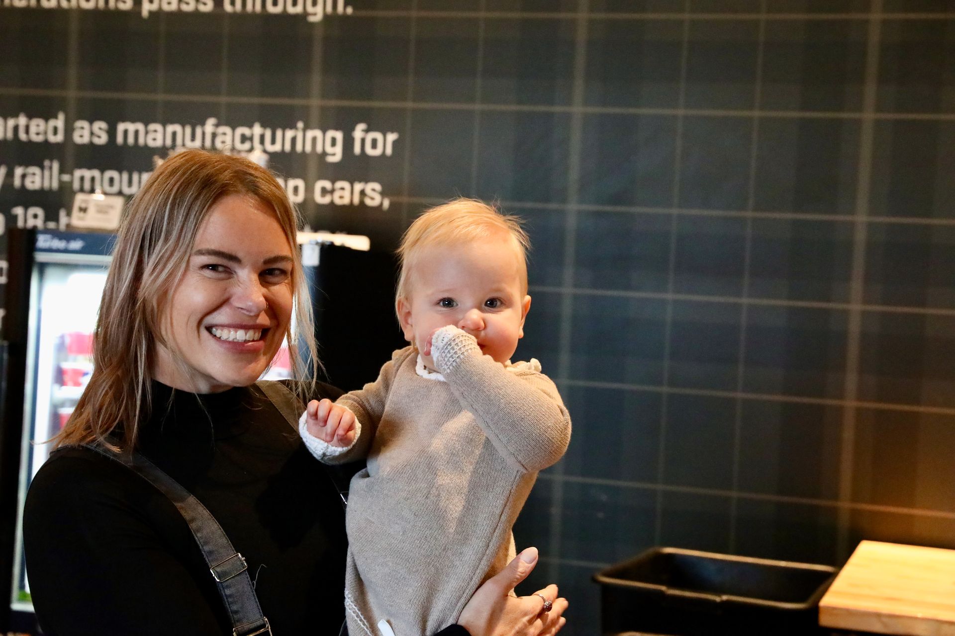 Woman holding a baby, smiling. Baby has hand in mouth. In front of a wall with text.