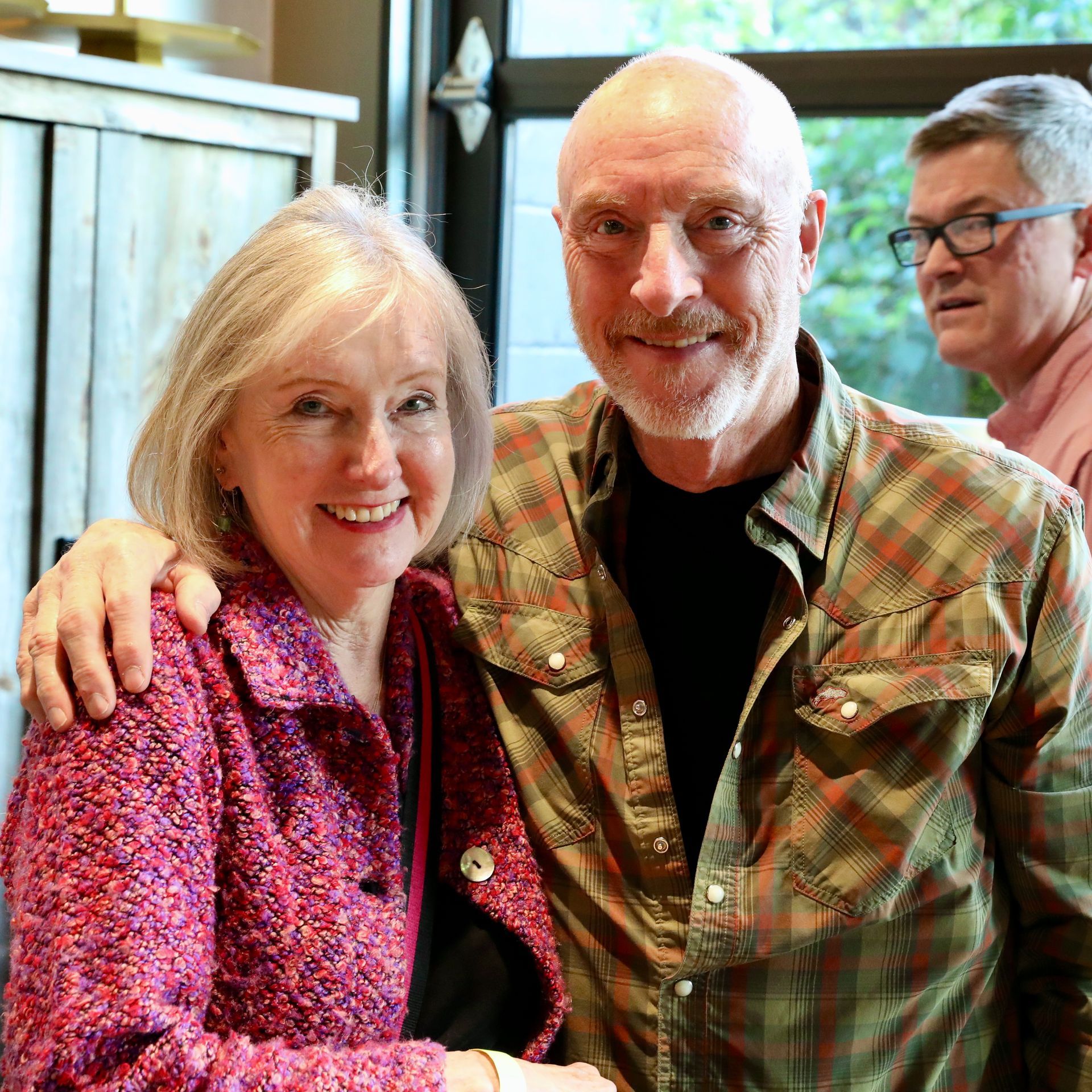 Woman in pink jacket and man in plaid shirt smiling, posing together, another man in background.