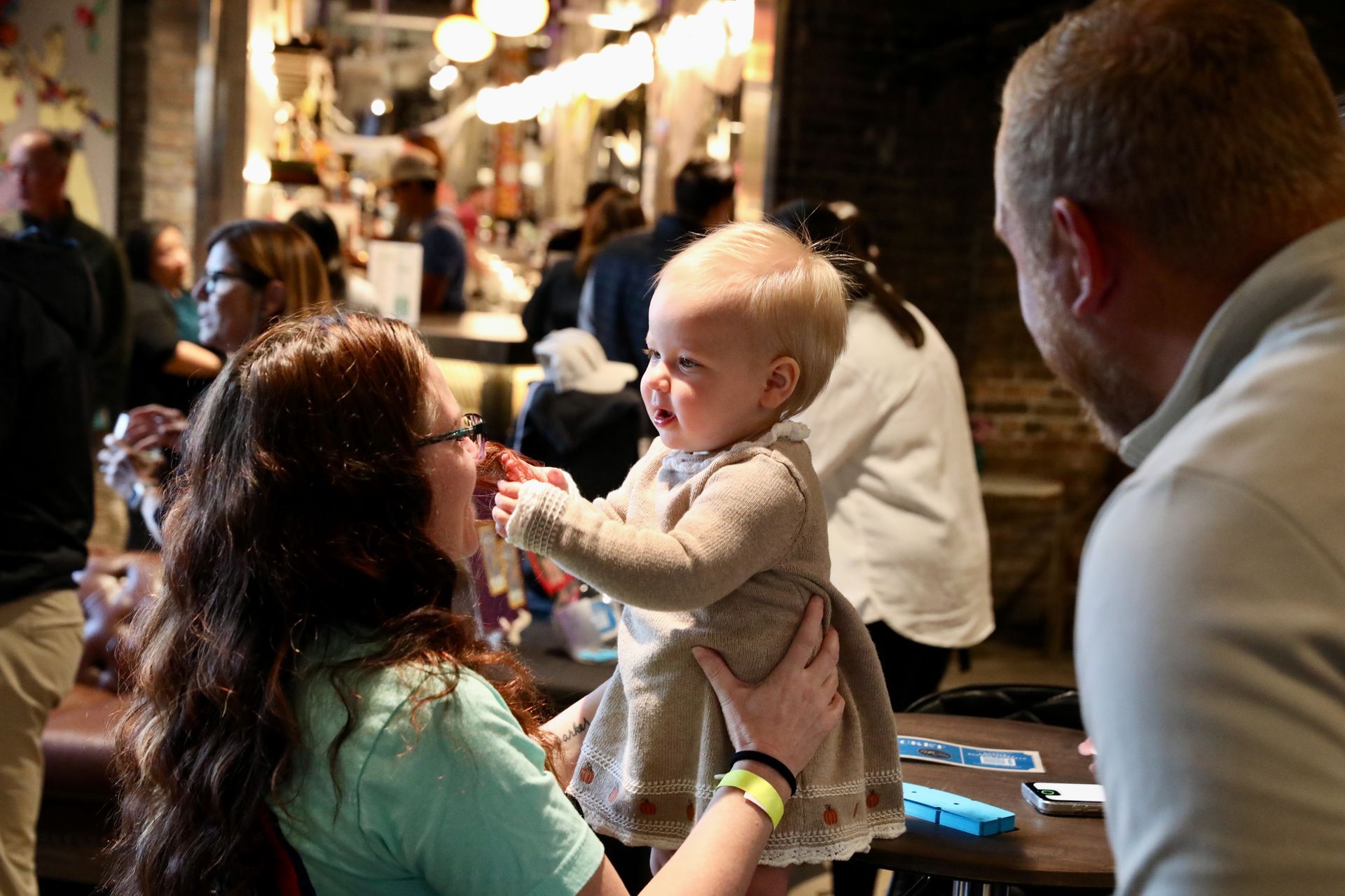 Baby reaching for something held by a person while another person looks on in a busy indoor setting.