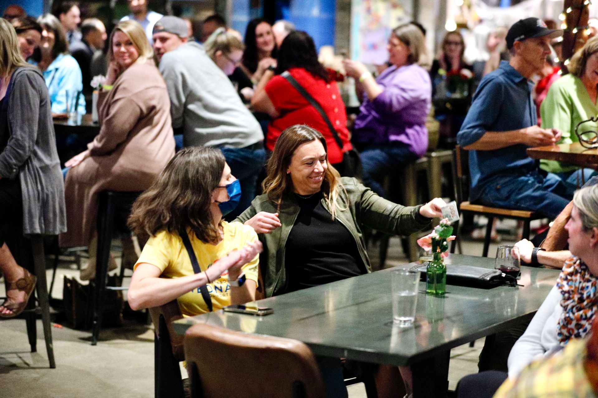 People at tables in a bar, chatting, holding drinks. Some seated on stools.
