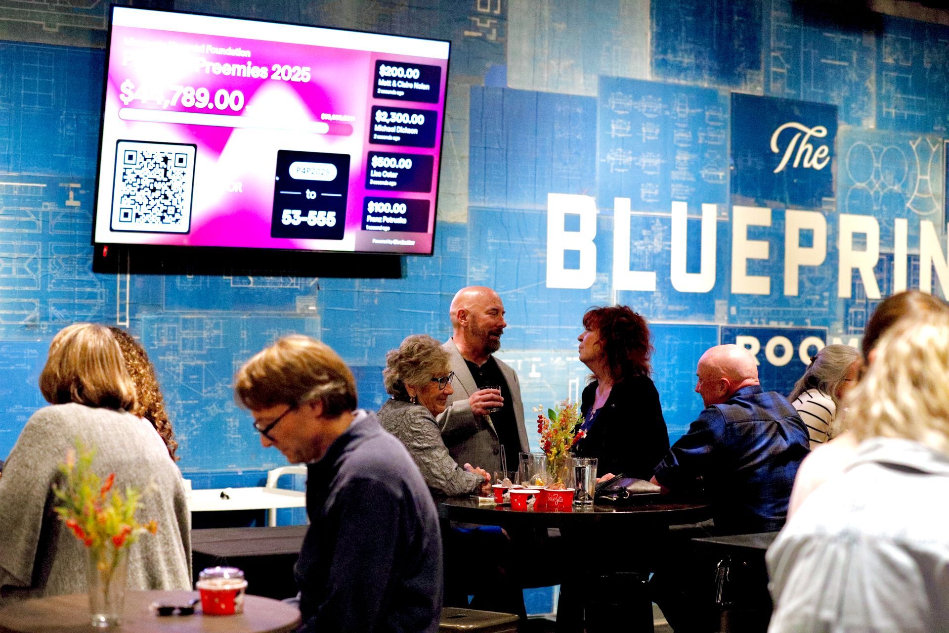 People at an event in a room with blue blueprint-themed walls. A screen displays numbers and a QR code.