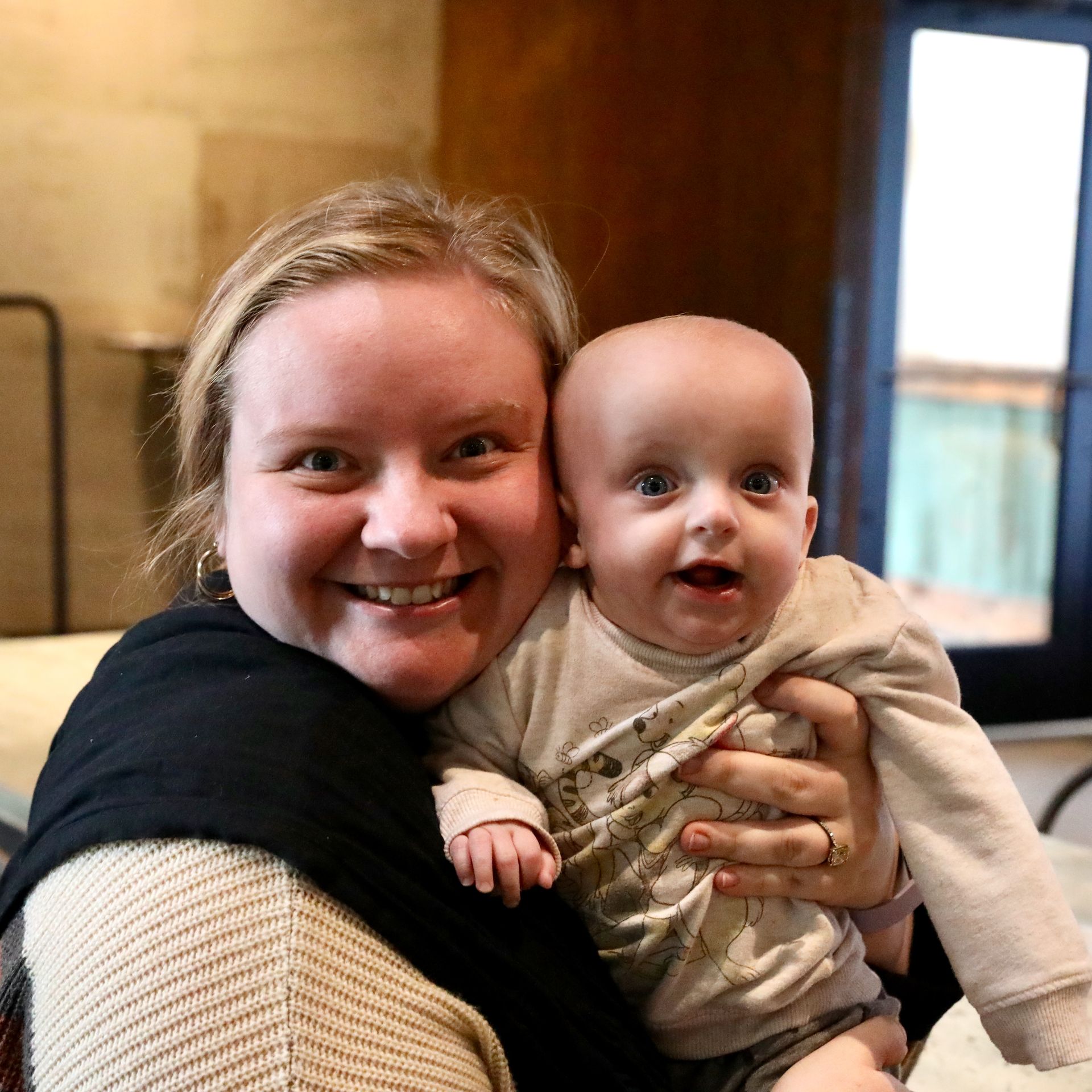Woman with blonde hair smiling, holding a baby. Both look at the camera. Neutral background.