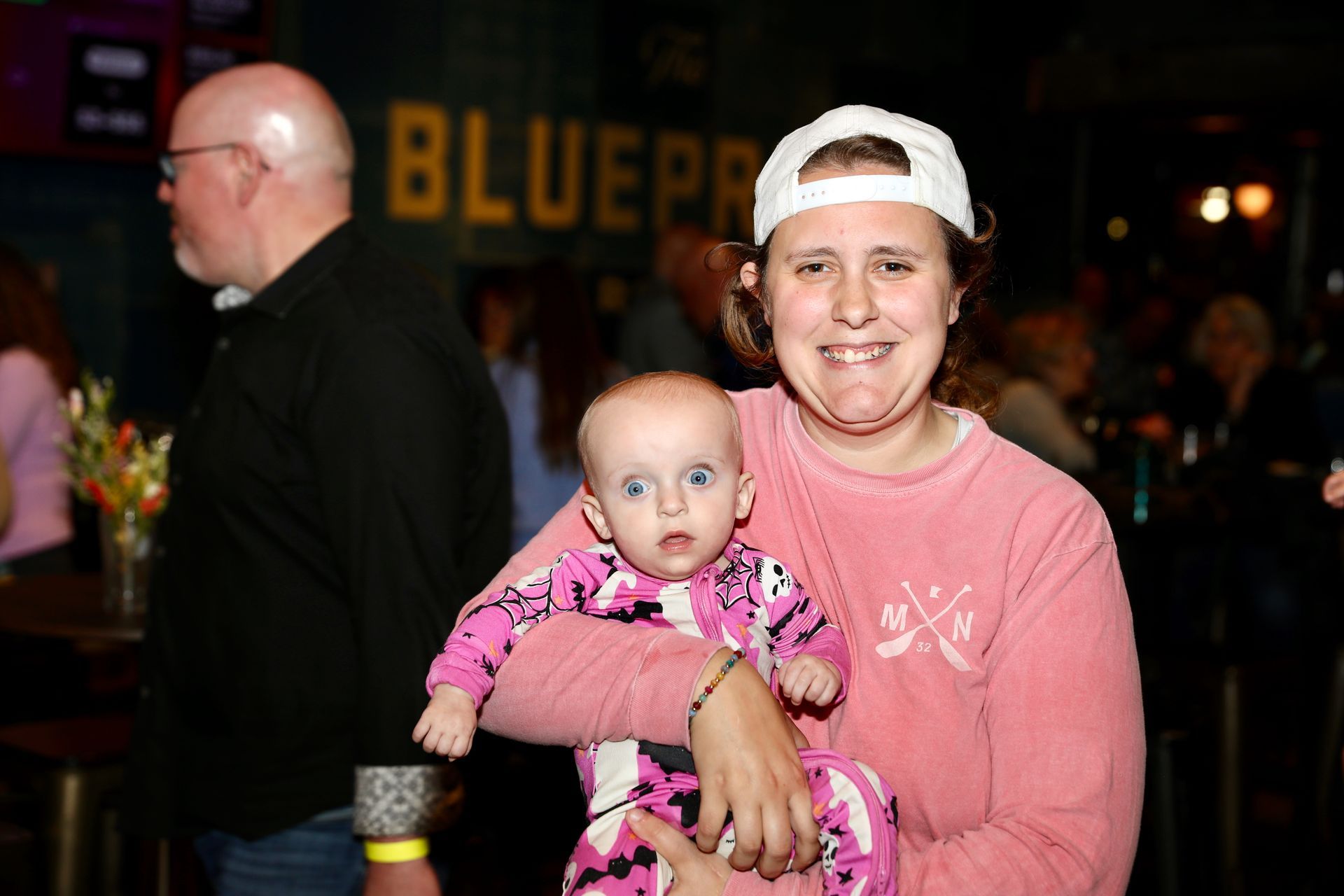 Woman in pink shirt and white cap holds baby in pink jumpsuit, smiling. Bar setting with people in background.
