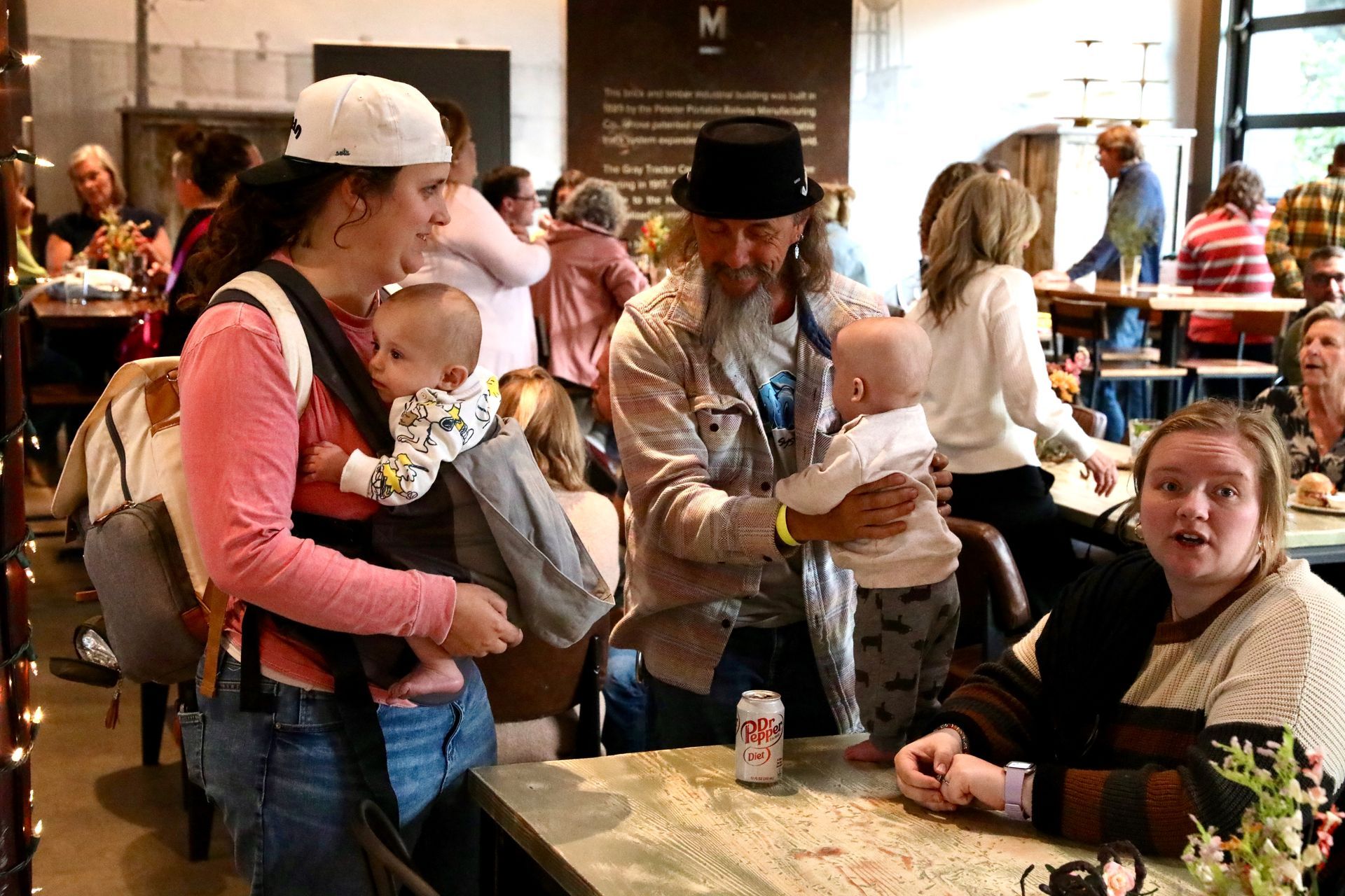 People with babies at a restaurant; one mother carries her baby. A man holds a baby. Another person sits at a table.