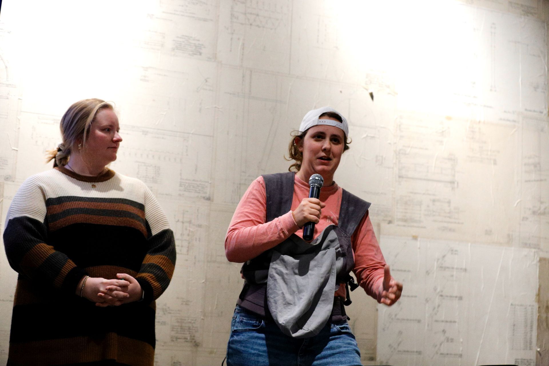 Two women on a stage, one with a microphone, speaking, wearing casual clothes. Background is a wall of paper.