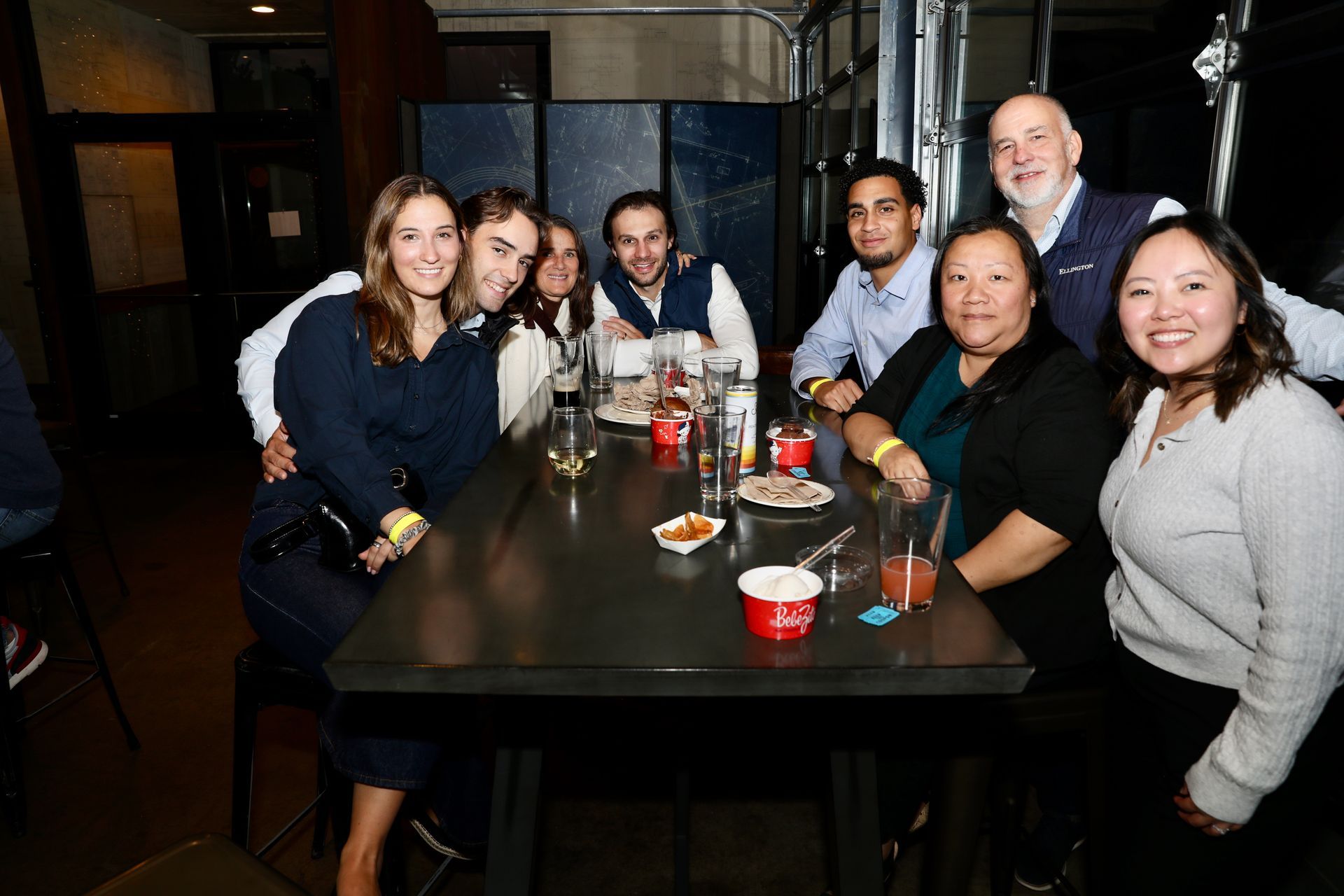 Group of people smiling at a table indoors. Dark setting with drinks and food.