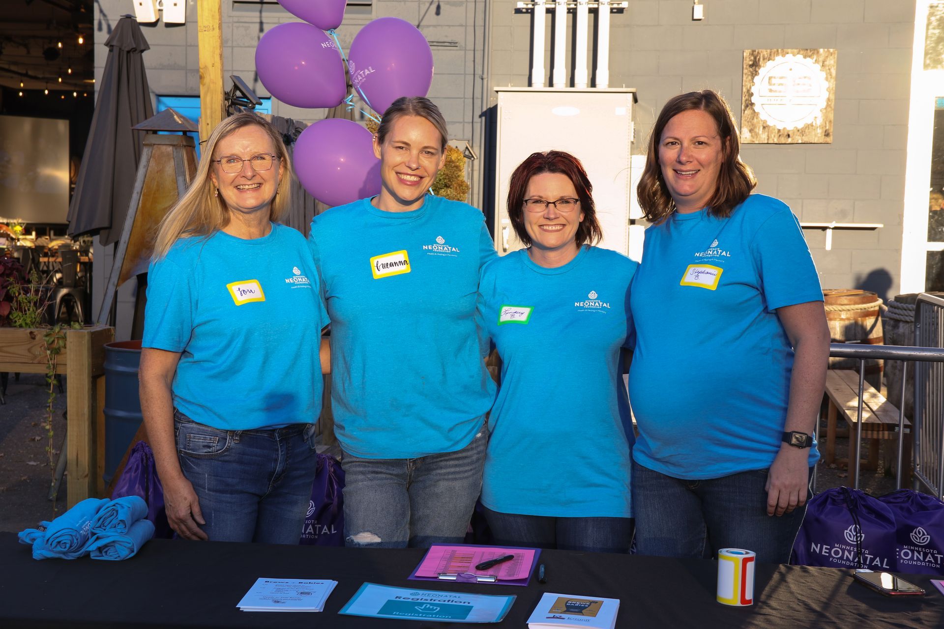 Four women in blue shirts stand behind a table with purple balloons. They are smiling.