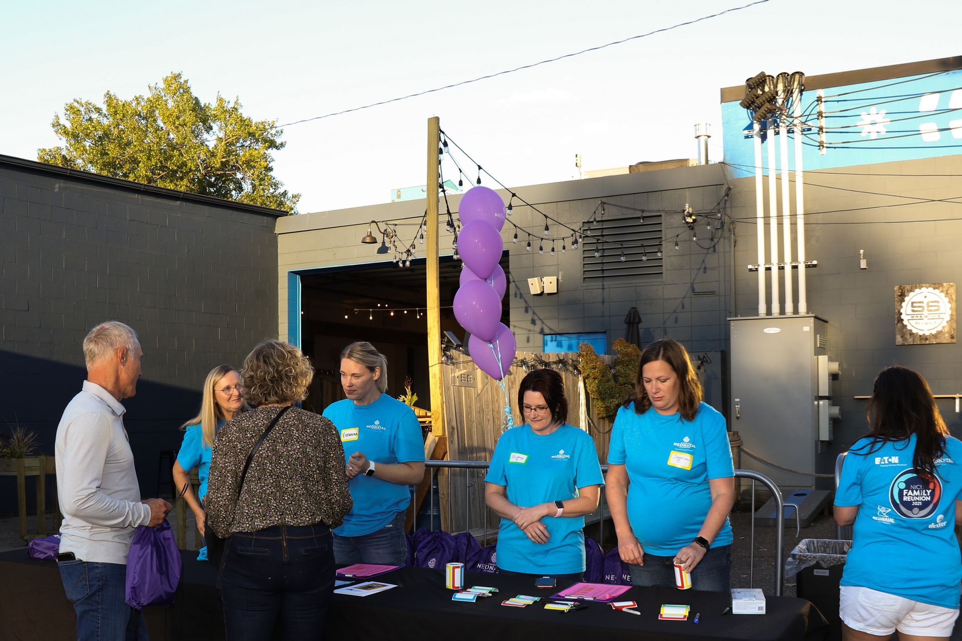 Volunteers at an outdoor event registration table, wearing teal shirts and assisting attendees. Purple balloons and decorations.