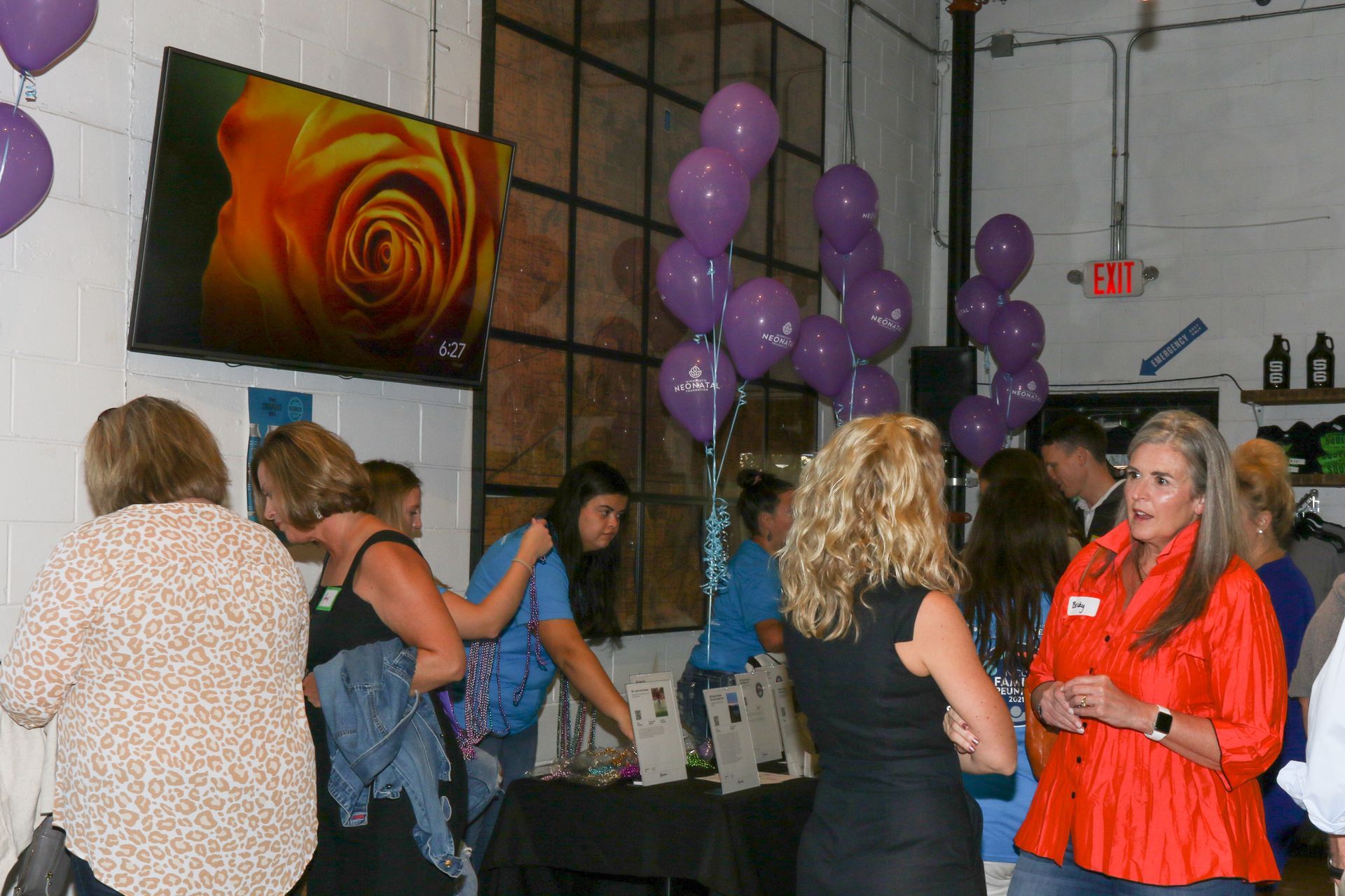 People at an event with purple balloons, a screen showing a rose, and a table with items.