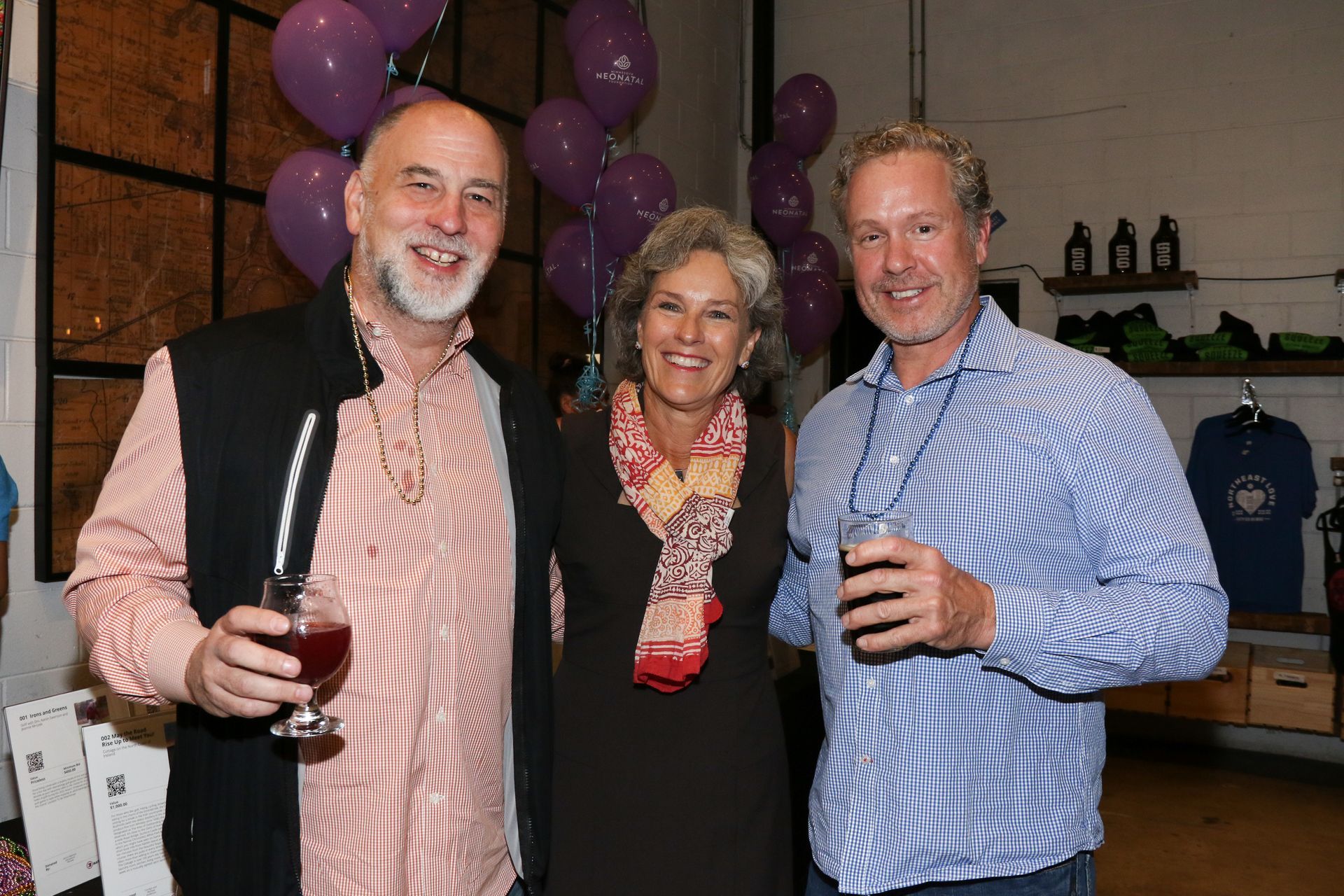 Three people smiling and holding drinks at an event with purple balloons and a brewery backdrop.
