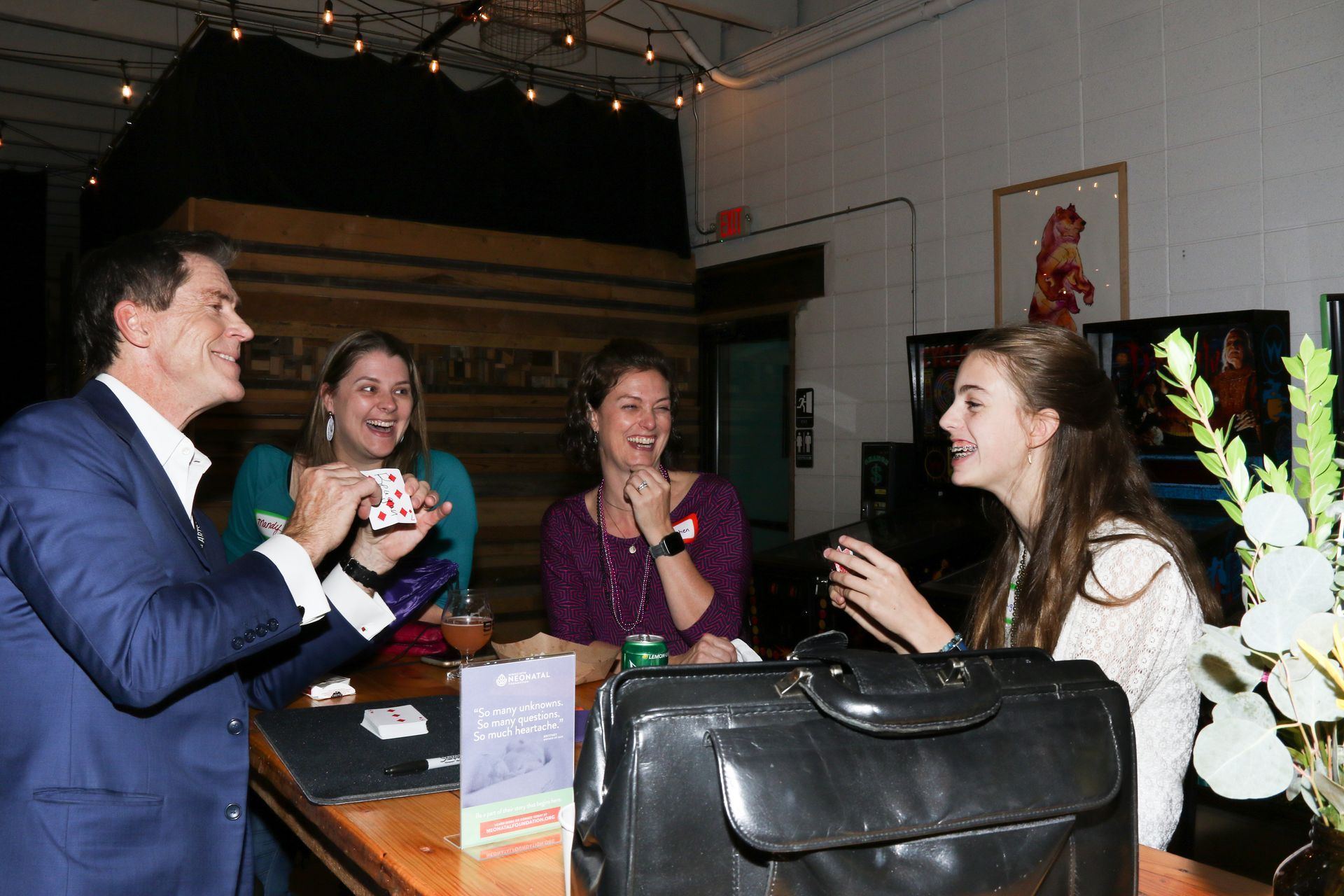 Man in suit performs card trick for three smiling women at a bar.