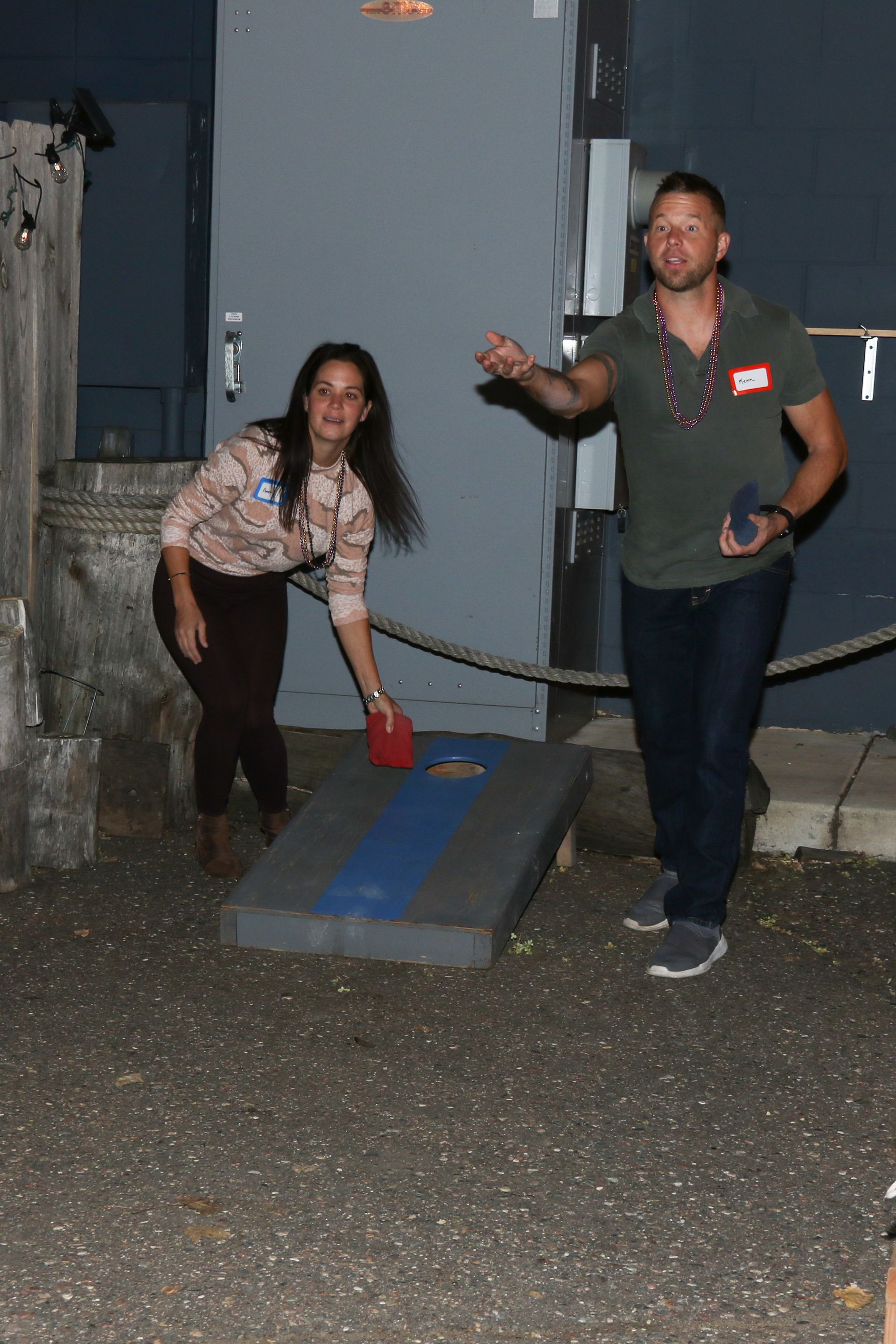 Two people playing cornhole outdoors at night; woman throws, man prepares to throw a beanbag.