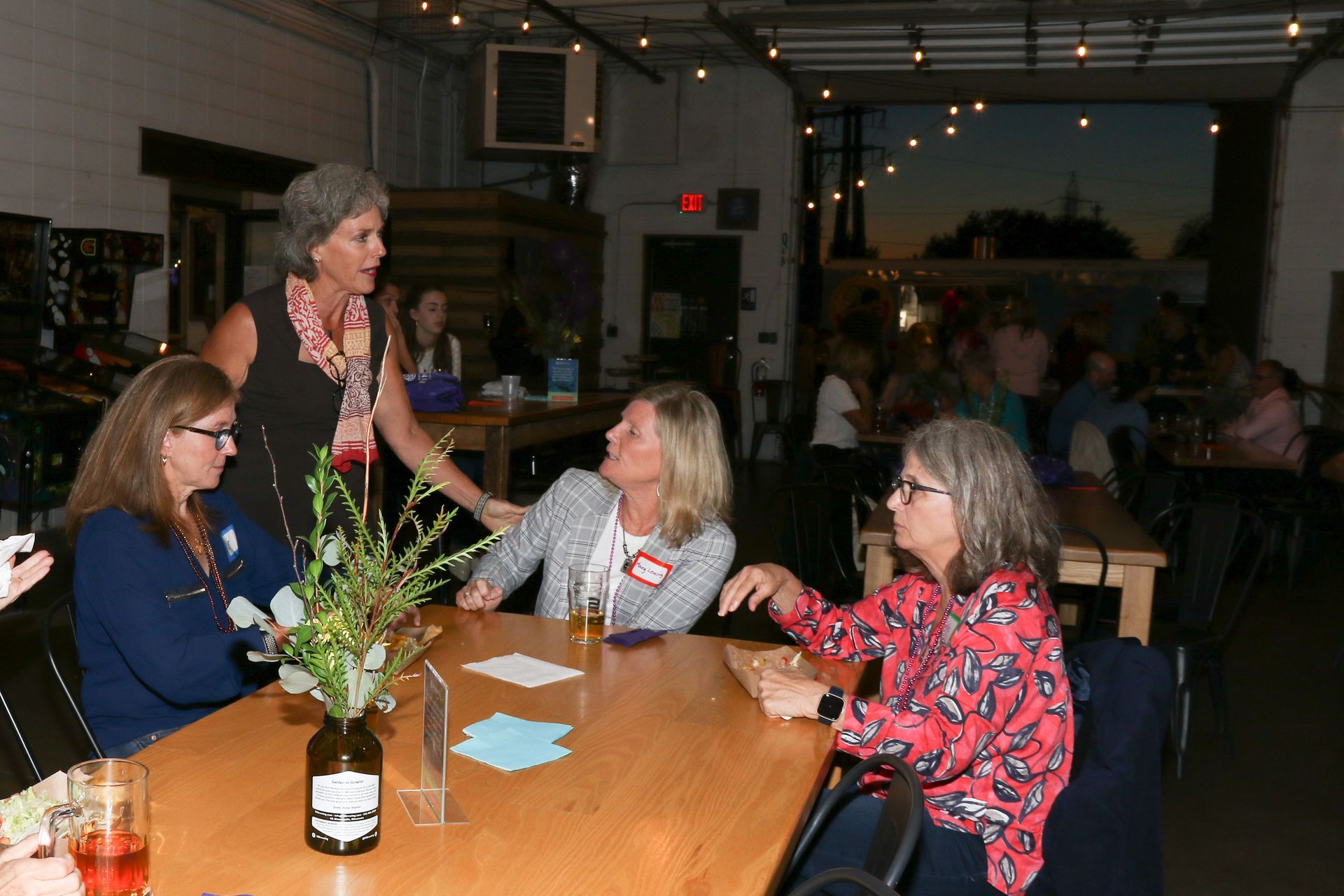 Four women at a table in a dimly lit event space; one woman gestures as she speaks, while others listen.
