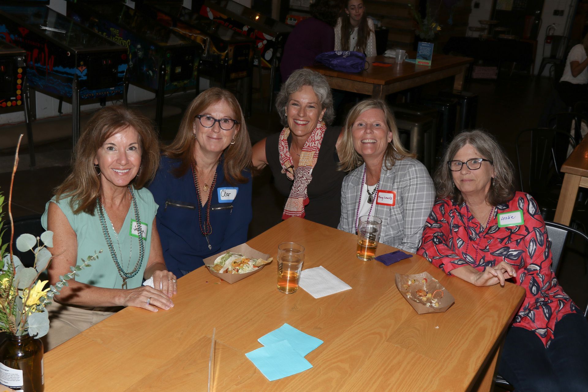 Five women smile at a table in a dimly lit room. Snacks and drinks are present.