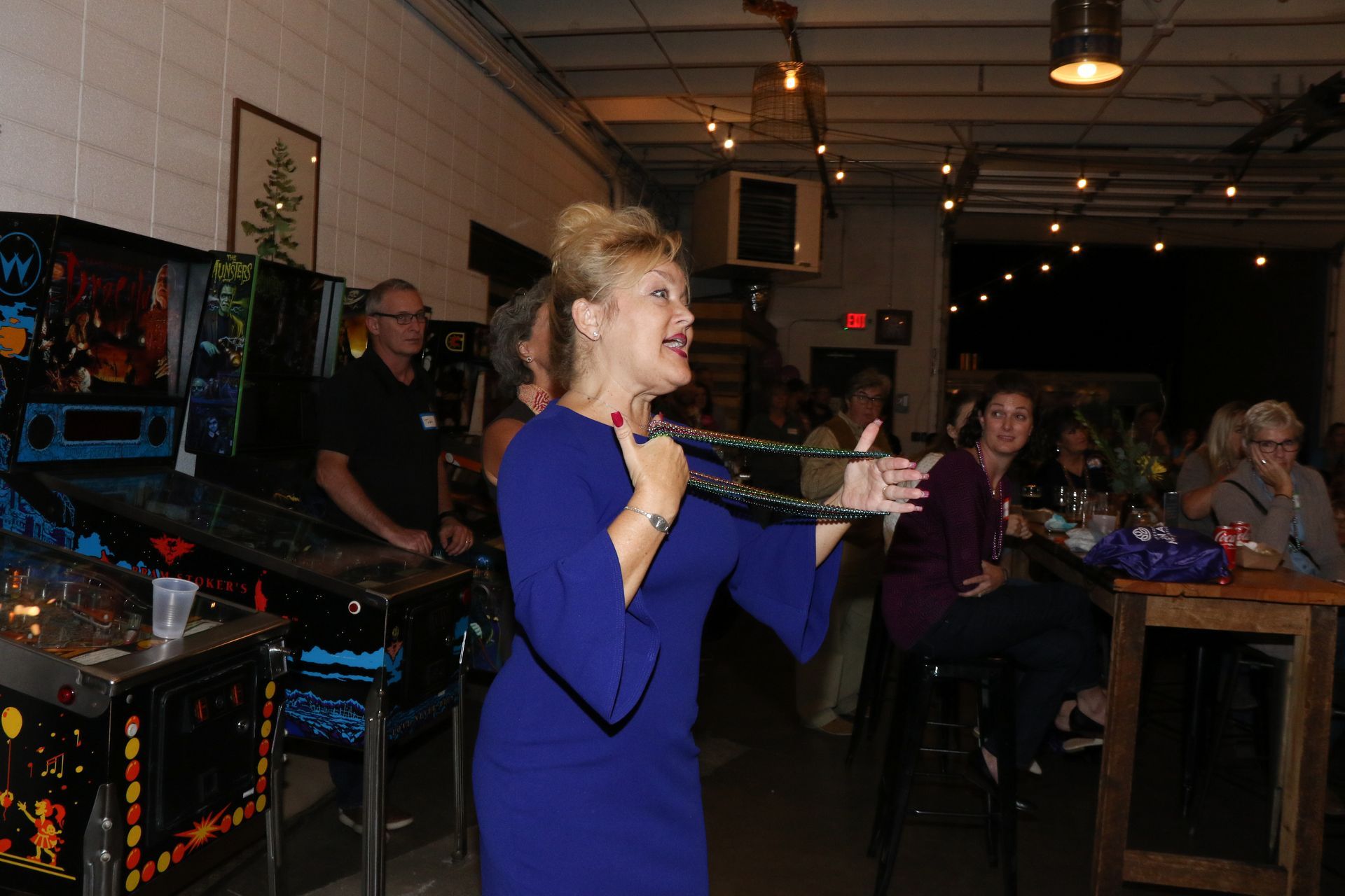 Woman in blue dress speaking, holding a cord, inside a bar with pinball machines and seated guests.