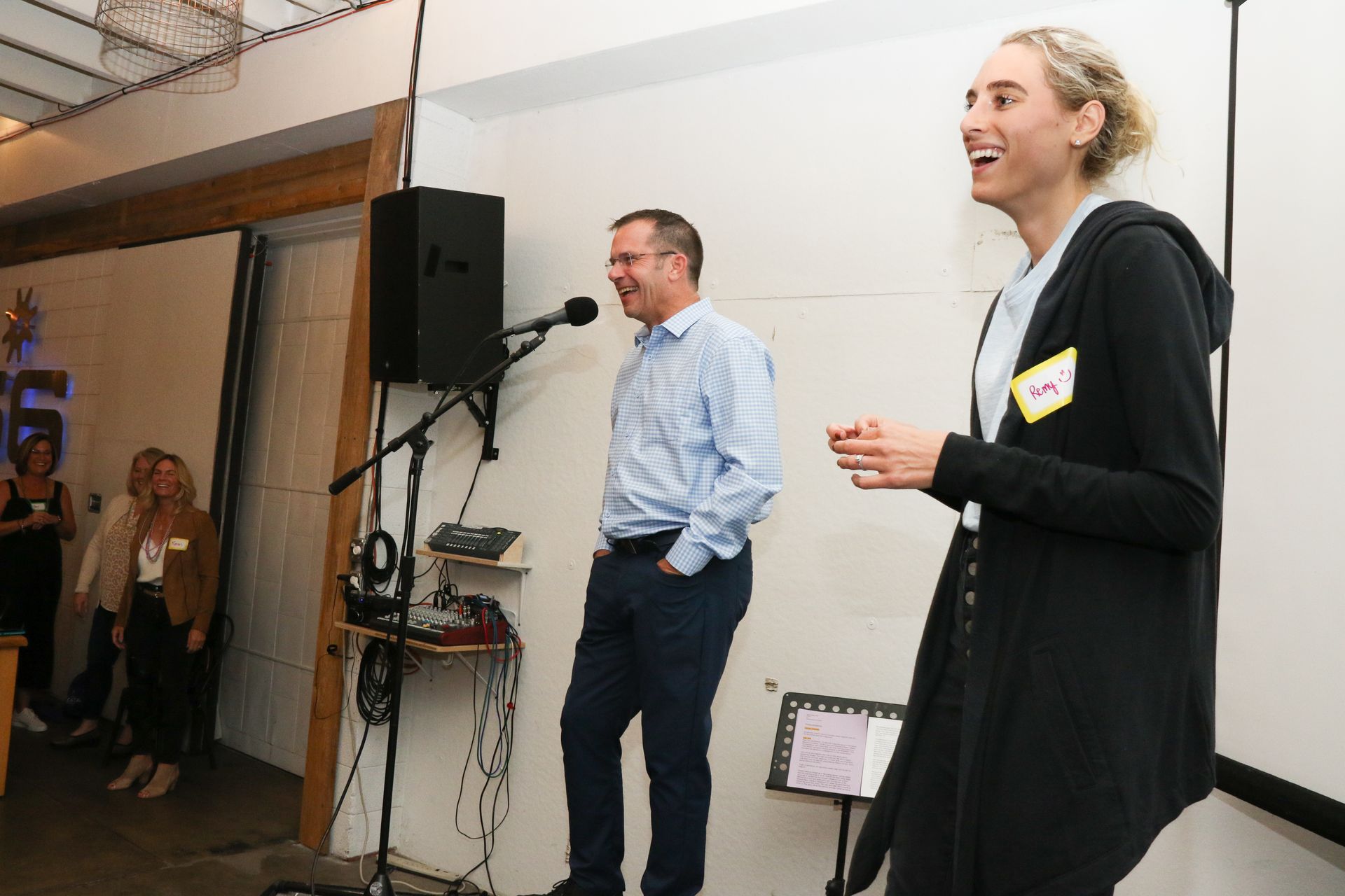 Two people on stage at an event: man speaking into a microphone, woman laughing, smiling.