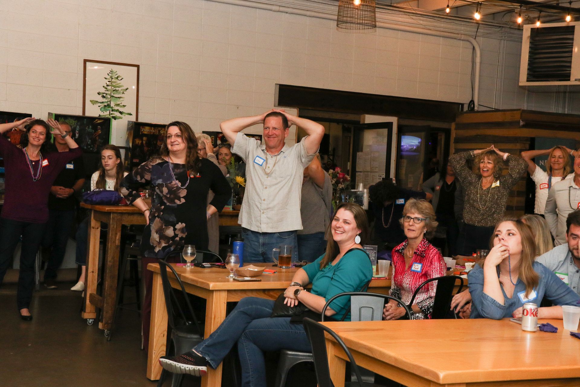 People in a room, some with hands on their heads, others seated. Tables and plants visible.