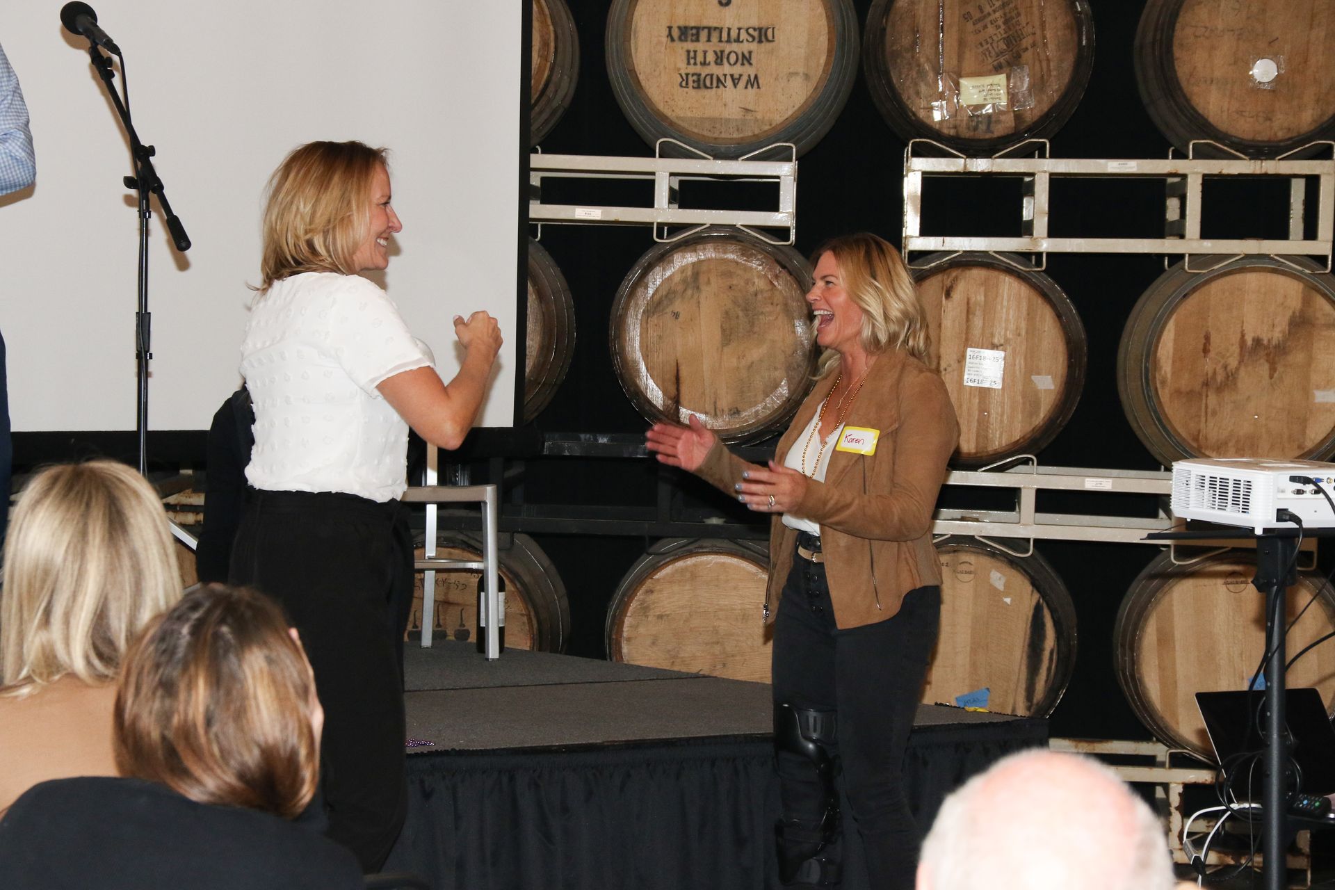 Two women laughing on a stage in front of barrels, speaking to an audience.