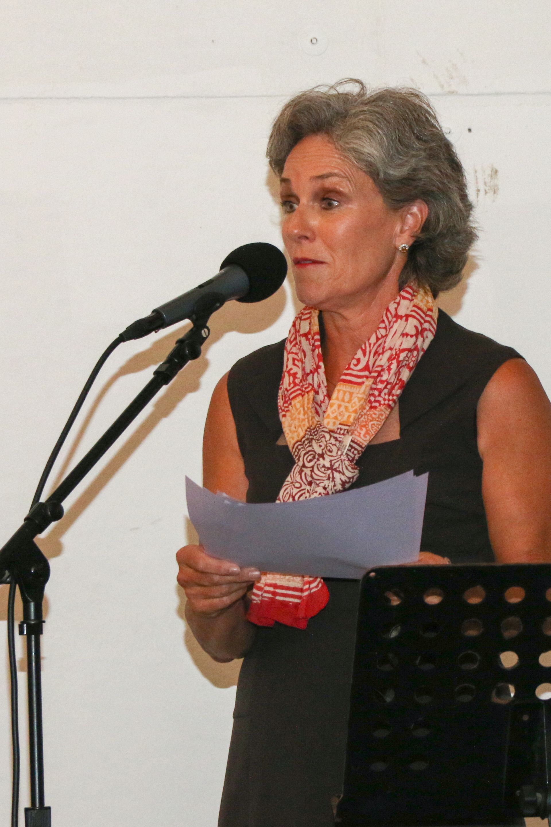 Woman speaking at a microphone, holding papers. She wears a patterned scarf and dark dress in a white room.