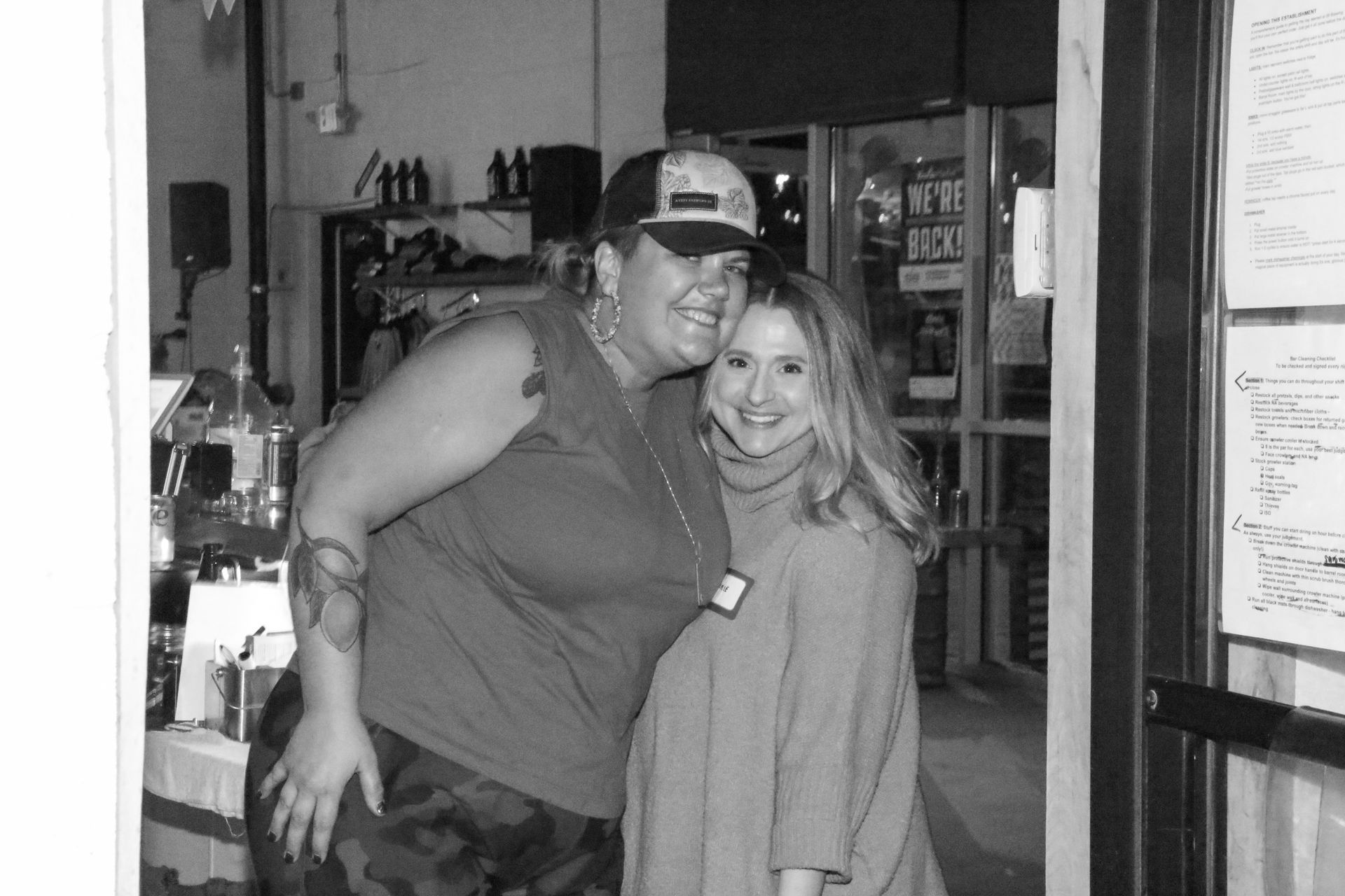 Two smiling people pose for a photo inside a bar. Woman with a hat and tattoo hugs a woman in a sweater.