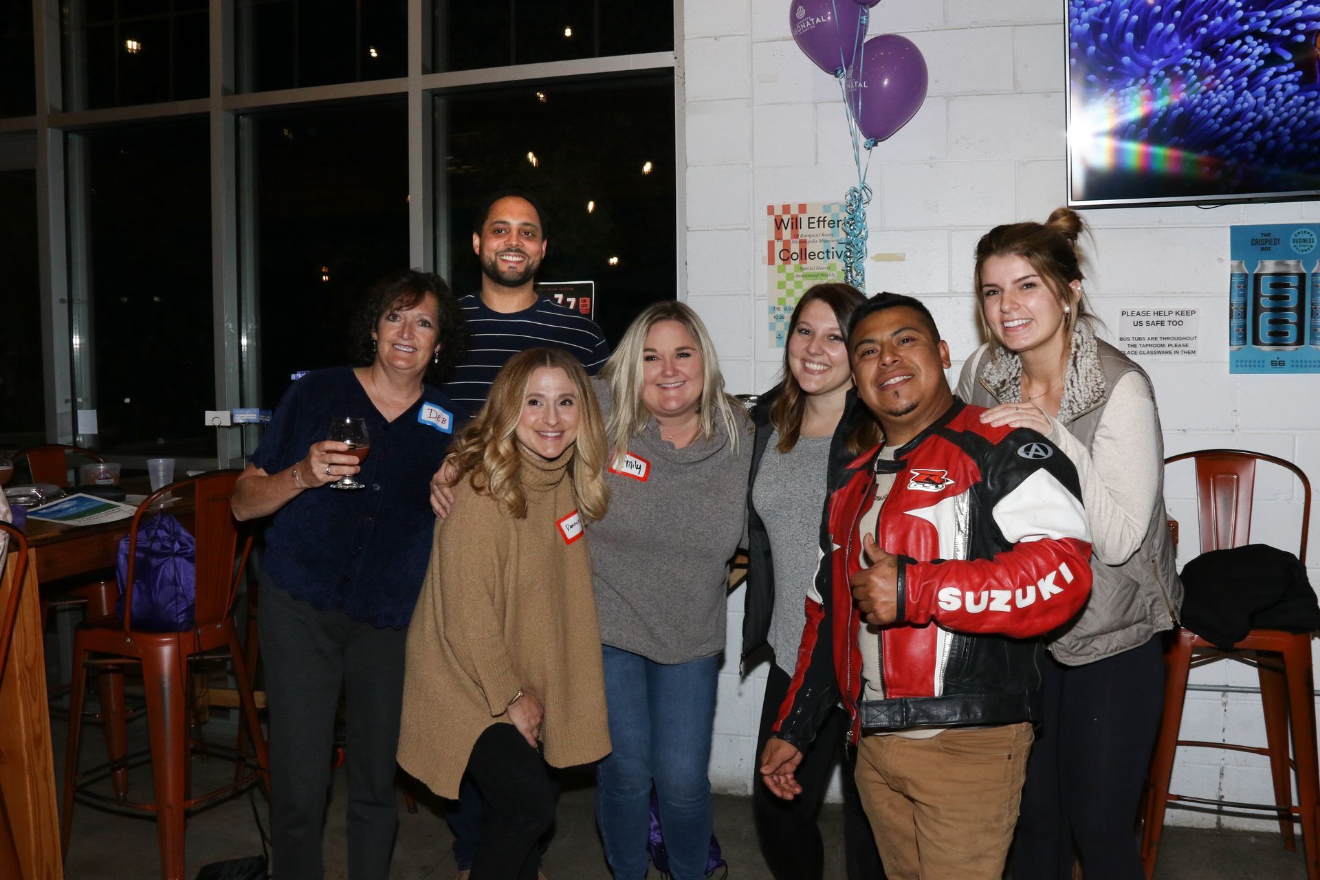 Group of people smiling at an event, near balloons and a bar, at night.