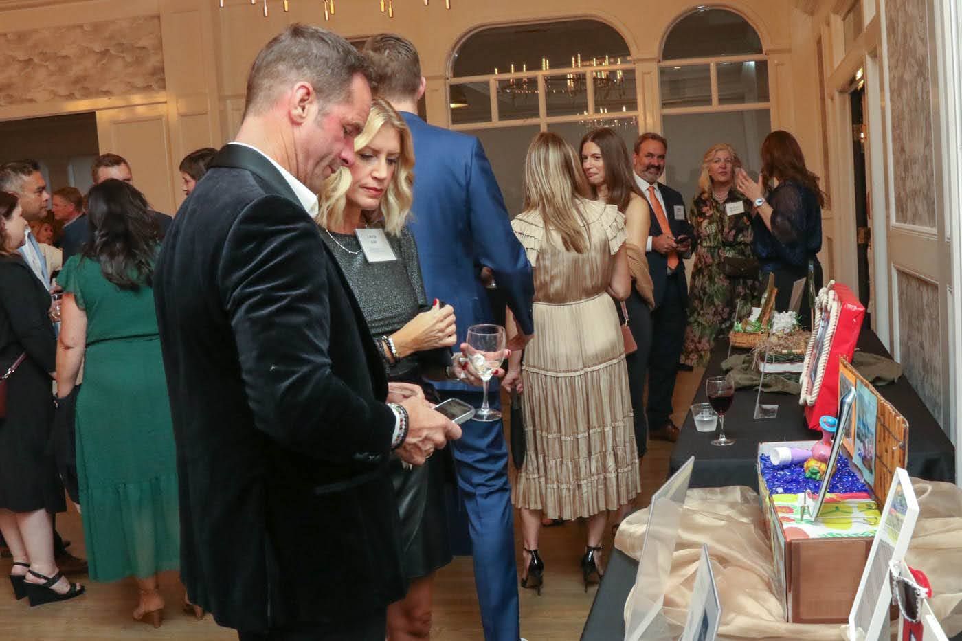 People socializing at an event with an auction table. A man in a velvet jacket looks at something.