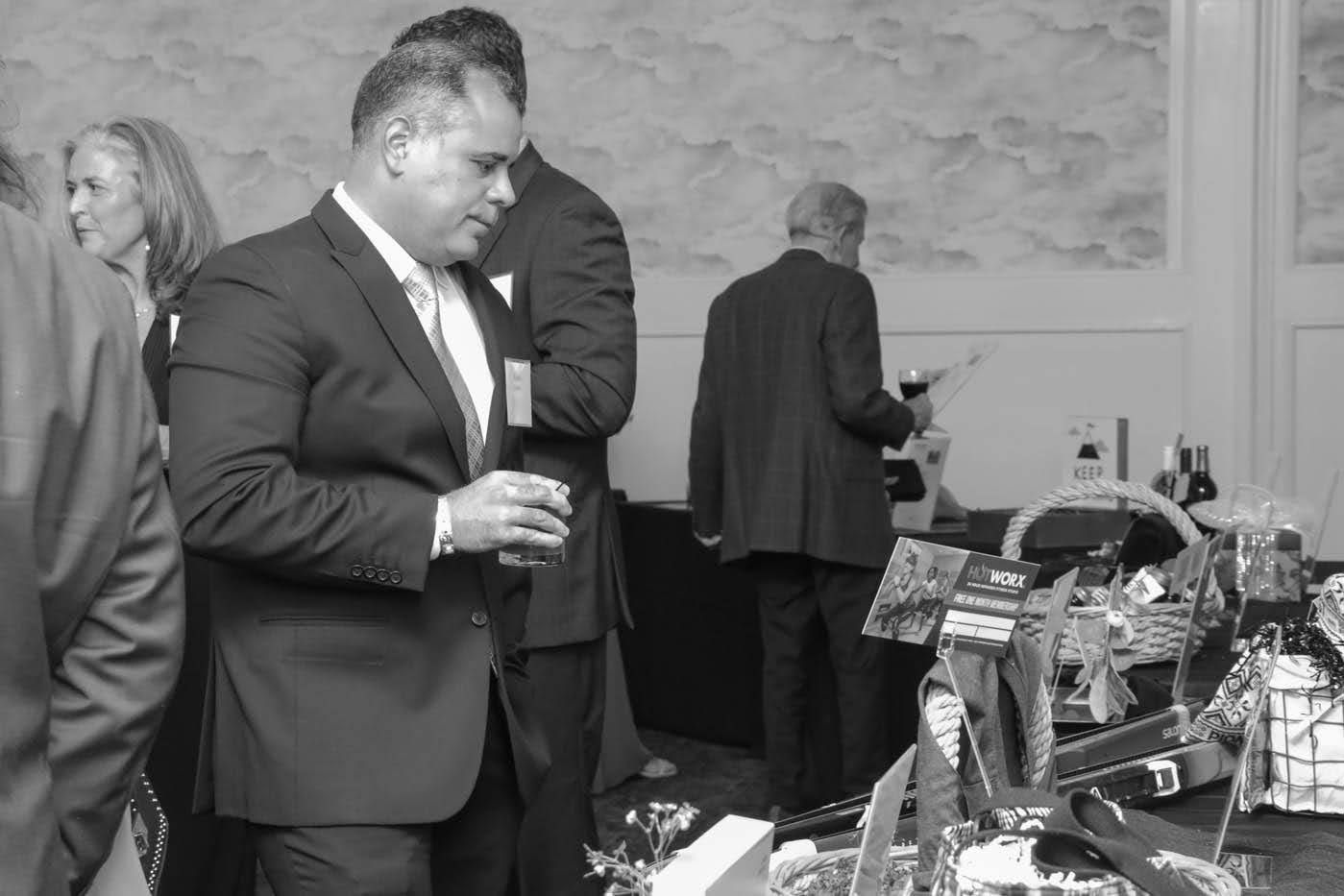Man in suit examines items on a table with other people in a room.
