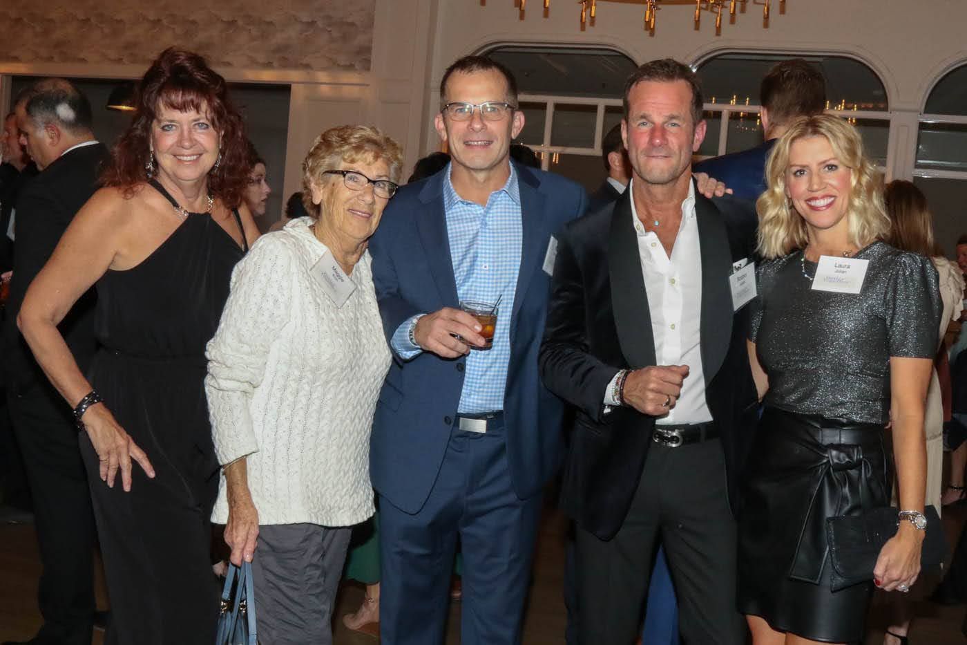 Five people at an event, smiling, posing for photo. Man in black tuxedo, others in semi-formal attire.