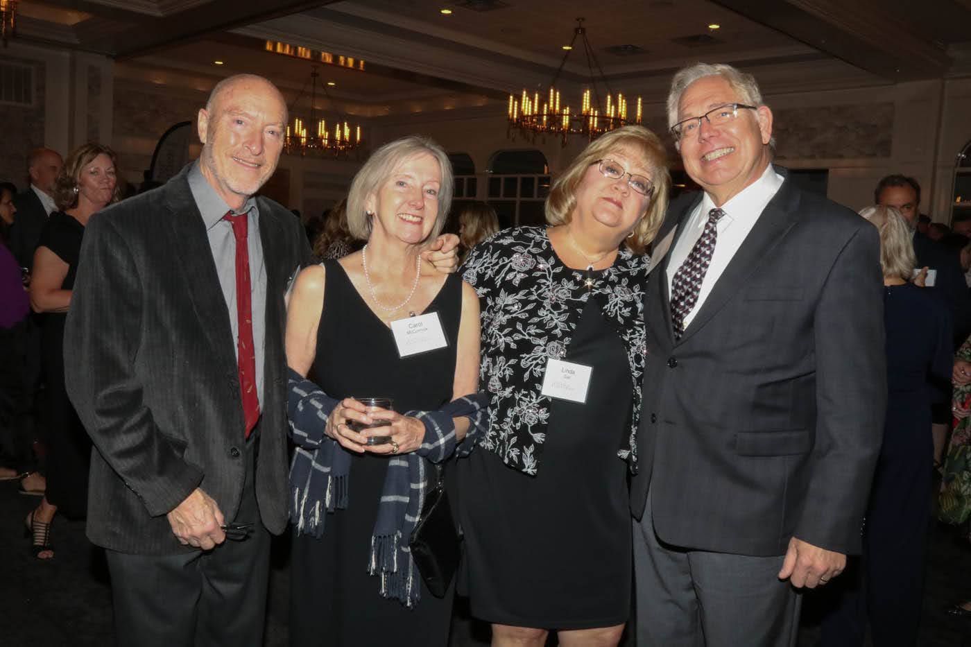 Four people smiling, posing together at an event. Formal attire, indoors.