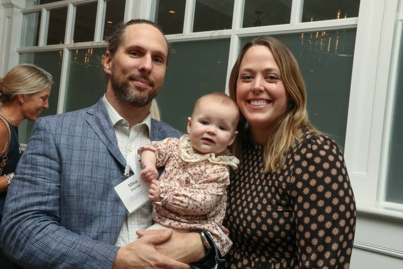 Man holding baby, smiling, with woman smiling beside them, standing in front of window.