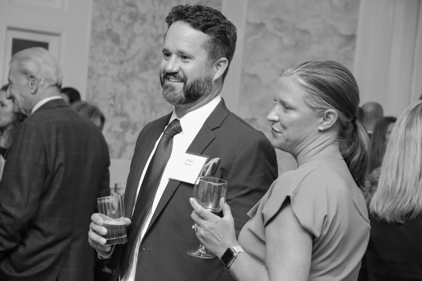 Man and woman at event, holding drinks, man wearing a suit, woman in a dress, conversing.