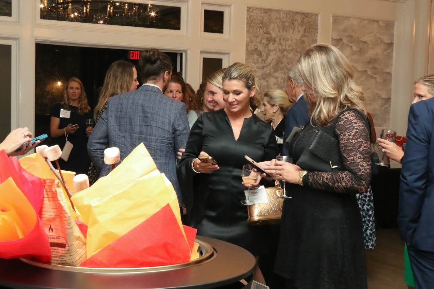 People at a gathering around a dessert station. One woman in black dress holds a phone and another holds a purse.