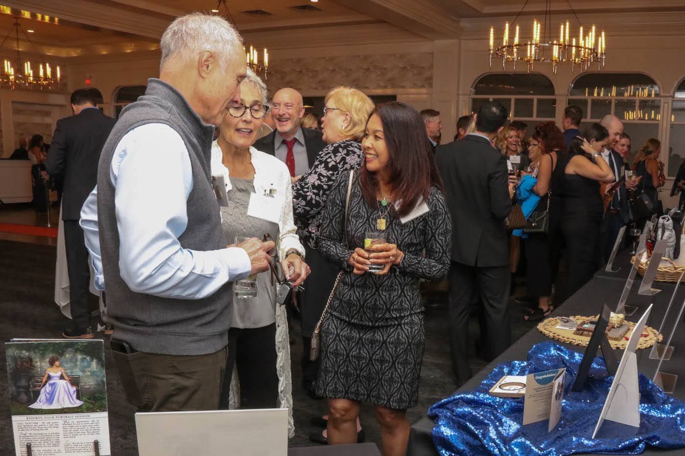 People chatting at an event, surrounded by tables with items. Dim lighting, formal setting.