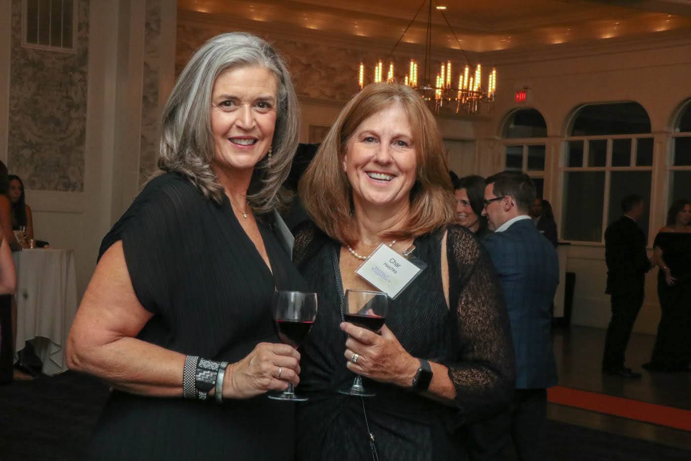 Two women smiling, holding wine glasses, at a formal event. One has gray hair, the other has brown hair.