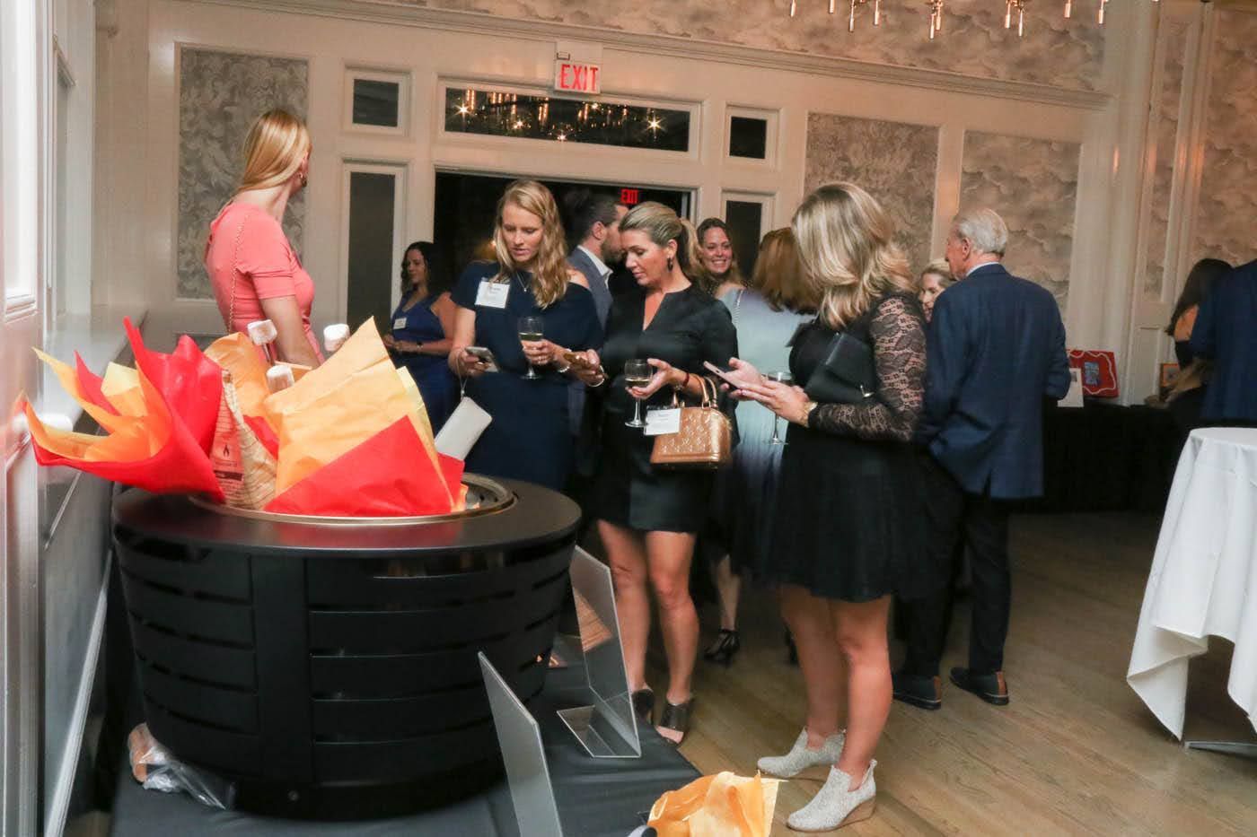 Guests at an event stand near a black fire pit and small tables in a well-lit room with ornate walls.