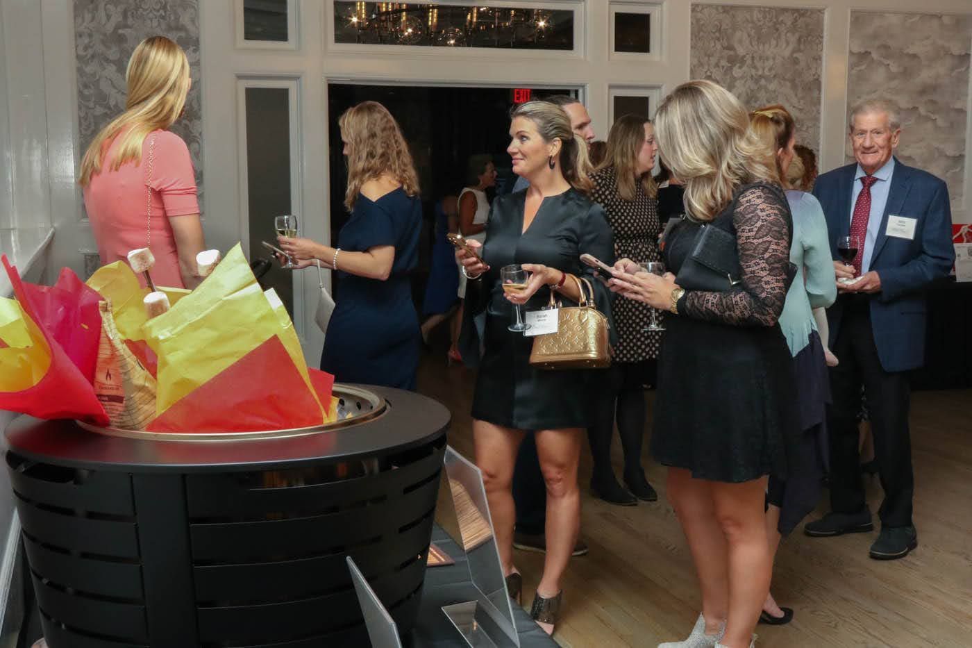 People at an event, some holding drinks, near a decorative display. A man in a suit and a woman with a gold purse are visible.