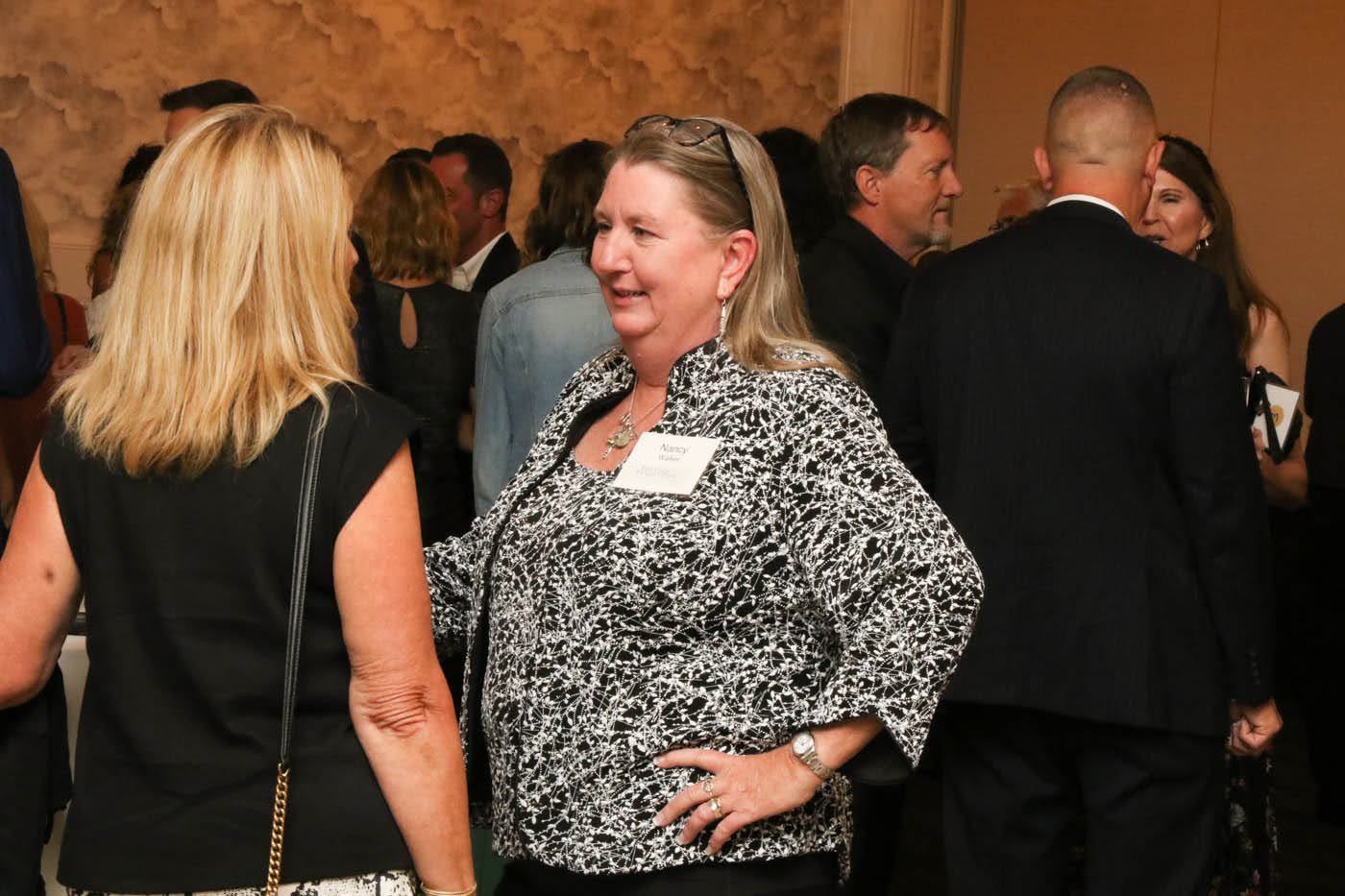 Woman with name tag in black and white patterned jacket chats with another woman at a social event.