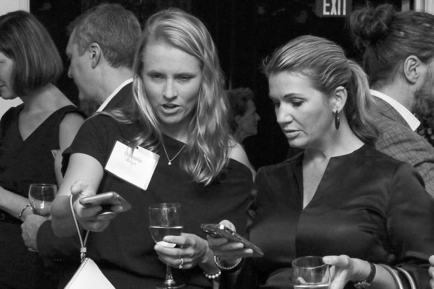 Two women looking at phones at an event, one holding a glass of wine, other people in the background.