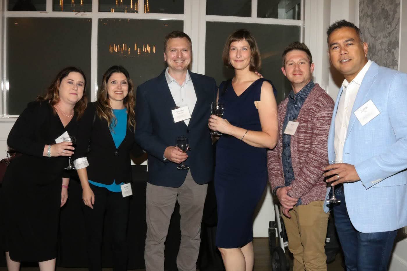 Six people at an event, posing together. A window in background. Some holding drinks.