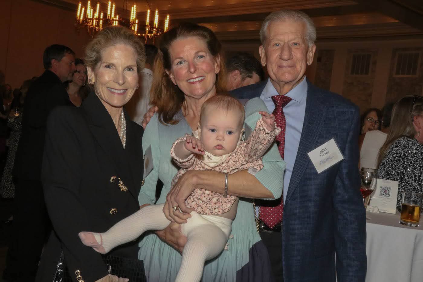 Group of four smiling people posing indoors. One holds a baby wearing floral.