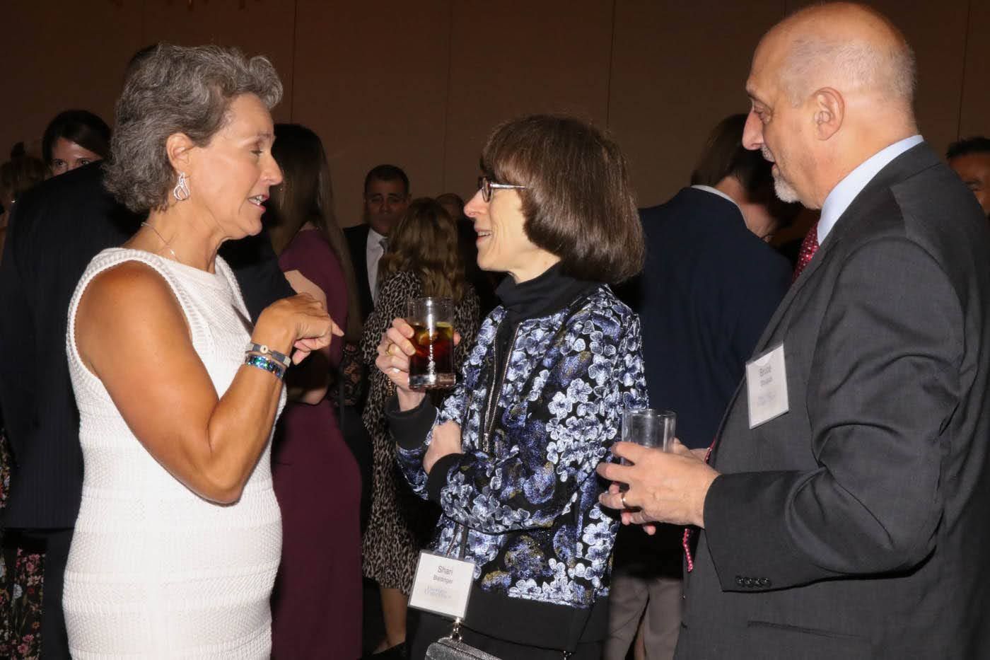 Three people conversing at a formal event. Woman in white dress gestures, woman in floral print holds a drink, man in suit listens.