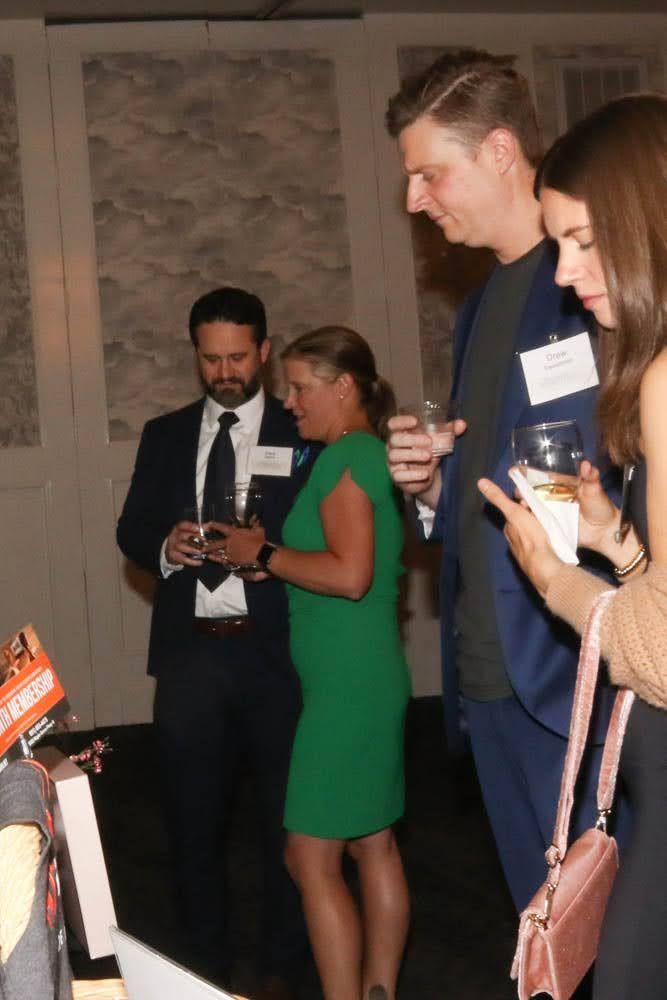 People at an event, some wearing name tags, holding drinks, and looking at something.